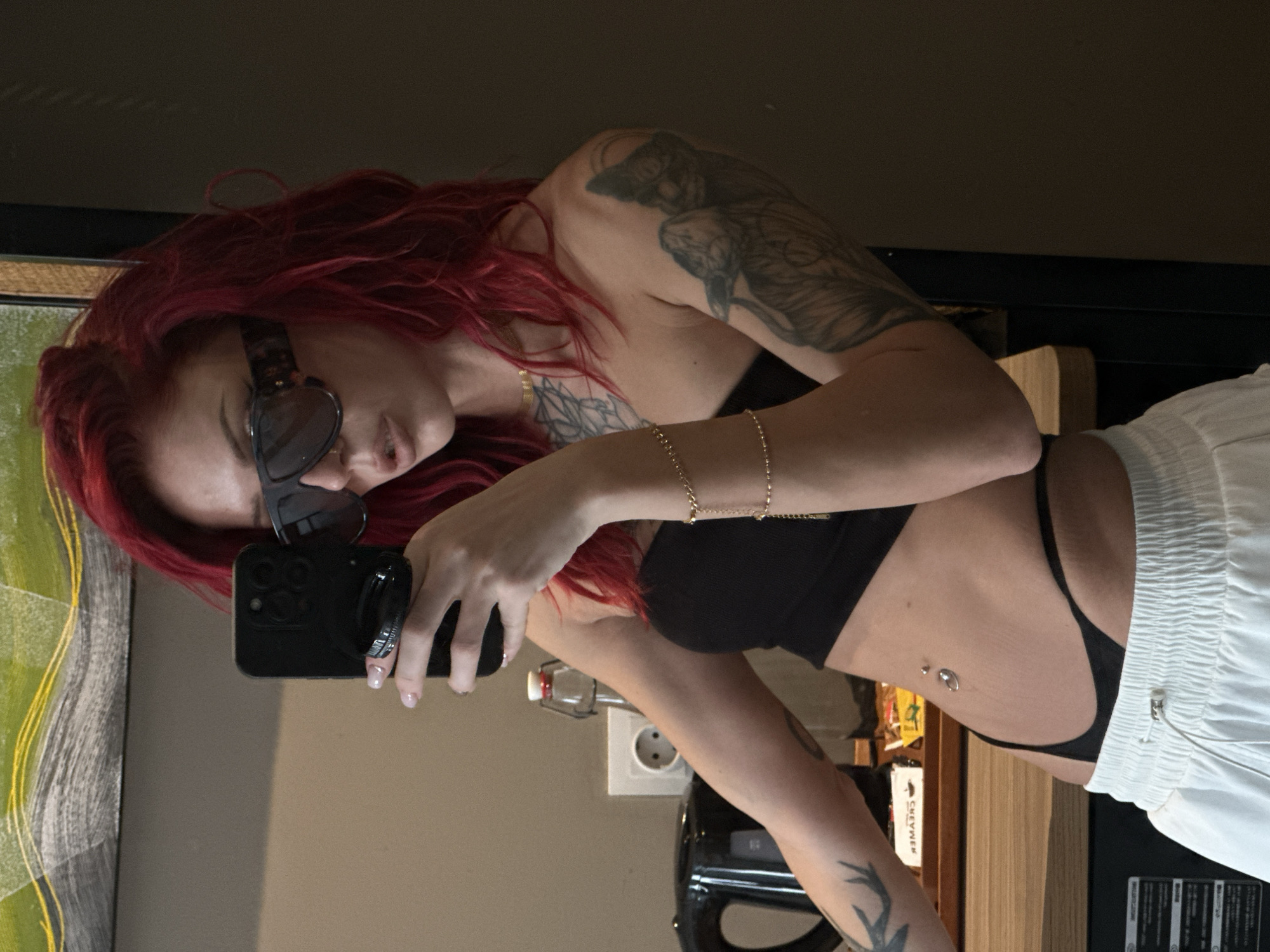 A woman with red hair, wearing sunglasses and a black bra, is taking a selfie in a room. Her skin has tattoos on her arm and chest. The background shows a wall, an electrical outlet, a painting, and a black kettle on a wooden surface. The location is in Unknown, Indonesia.