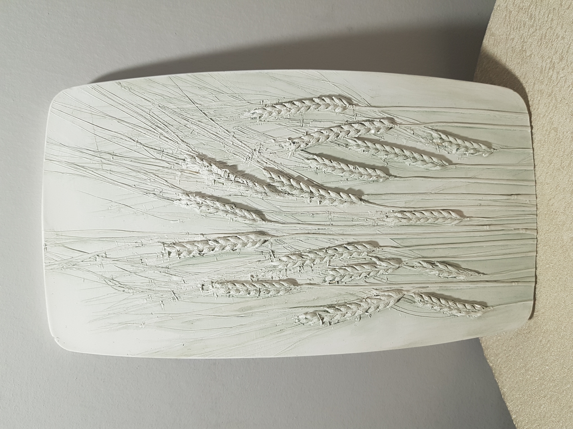 A rectangular, off-white ceramic tile with a raised relief depicting stalks of wheat sits against a light gray wall and a beige patterned wall. The tile has rounded corners and a matte finish. The wheat stalks are arranged in two staggered rows, with several stalks in each row. Fine lines are etched into the surface of the tile to represent the wheat stalks' stems and leaves. The tile is partially in shadow. The image evokes a sense of rustic simplicity and craftsmanship. There is no indication of this image being in Saint Petersburg, Russia.