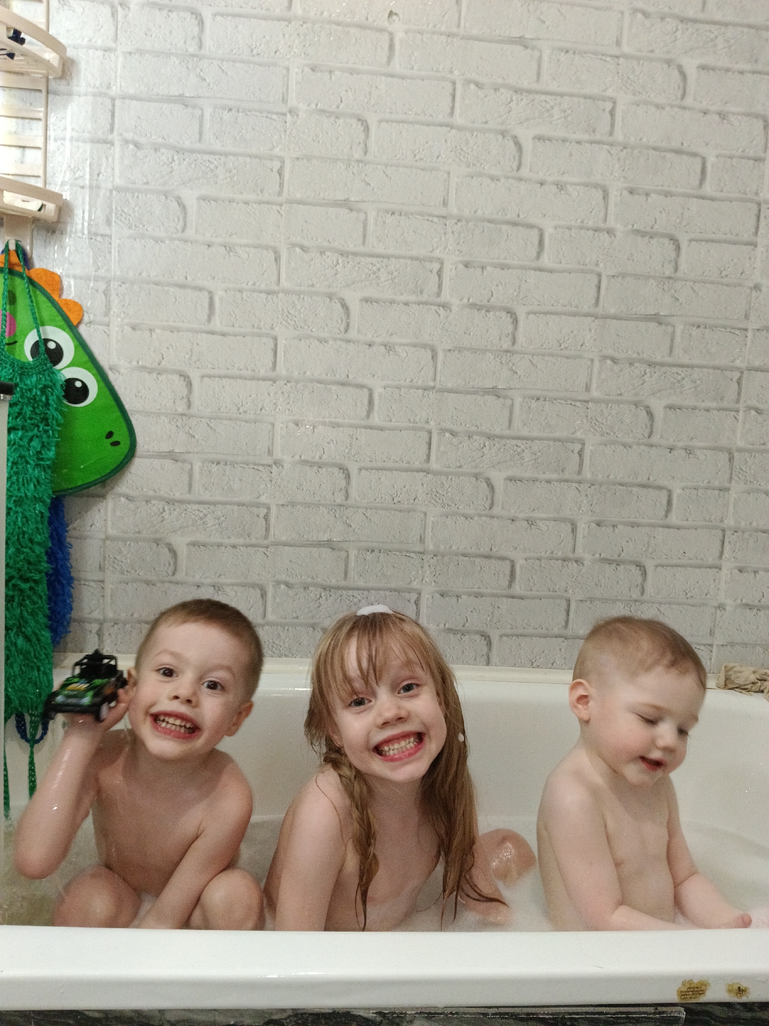 Three children are seated in a bathtub filled with water and bubbles. The child on the left is a boy with short reddish-brown hair and a toy car in his left hand; he is grinning. The child in the center is a girl with long, wet reddish-blonde hair. She is also smiling and has a small mound of foam on her head. The child on the right is a younger boy with short reddish-blonde hair; his head is slightly bowed and he seems to be looking downward.
The background consists of a white brick-patterned wall. To the left of the children, a green dinosaur-shaped bath toy organizer hangs on the wall.
The location is a bathroom, presumably in a private residence. There is no information in the image to confirm this is in Chaikovsky, Russia.