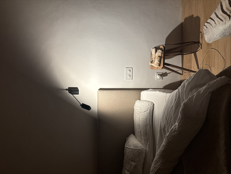 The image shows a bedroom in Celerina/Schlarigna, Switzerland. The bed is made with white linens and a brown headboard, with a white throw blanket draped over the top of the bed. A small wooden stool sits next to the bed, with a white fur throw on top. A wall-mounted lamp with a black cord is casting a light onto the bed. The wall is white with a light switch near the bed. The floor is made of wood. The environment is quiet and peaceful. There are no people or other animals present in the image.