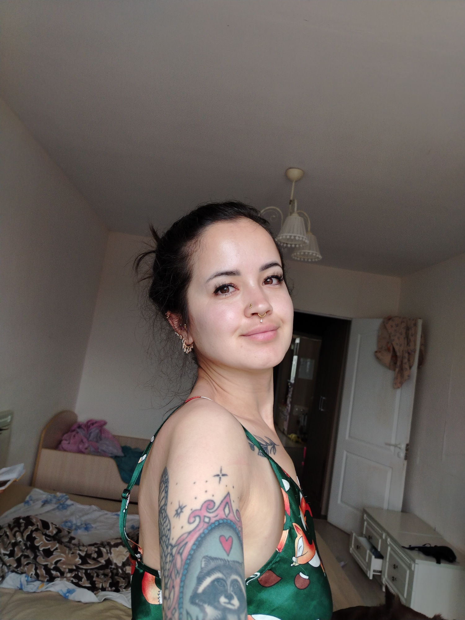 The image shows a woman with dark hair pulled back in a bun, wearing a green slip dress with a cartoon print. She has tattoos on her left arm and shoulder, including a raccoon portrait. She is wearing earrings and a nose ring. The woman is posing for a selfie in a bedroom. The room has a bed with various bedding, a bedside table, a wardrobe, and a dresser. The overall setting suggests a domestic environment. The photograph was taken indoors, likely in the city of Noviy, Russia, given the inferred location.