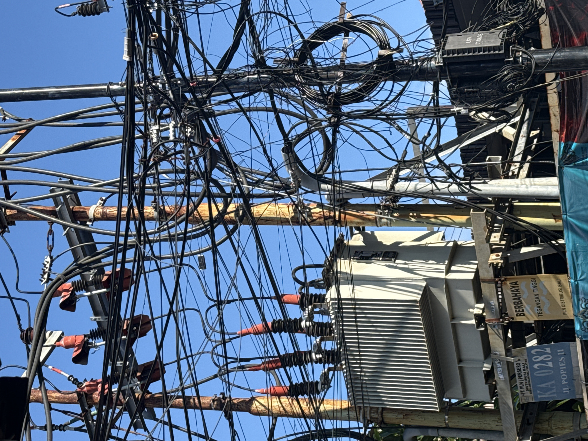 The image depicts a dense cluster of electrical wires and equipment mounted on utility poles against a clear blue sky. The location is Kuta, Indonesia, as indicated by the sign "JL POPPIES II" and "PLN Distribusi Bali" (State Electricity Company of Bali). There is a transformer with visible cooling fins and insulated electrical connections. Signs attached to the pole convey warnings about high voltage ("BERBAHAYA TEGANGAN TINGGI").