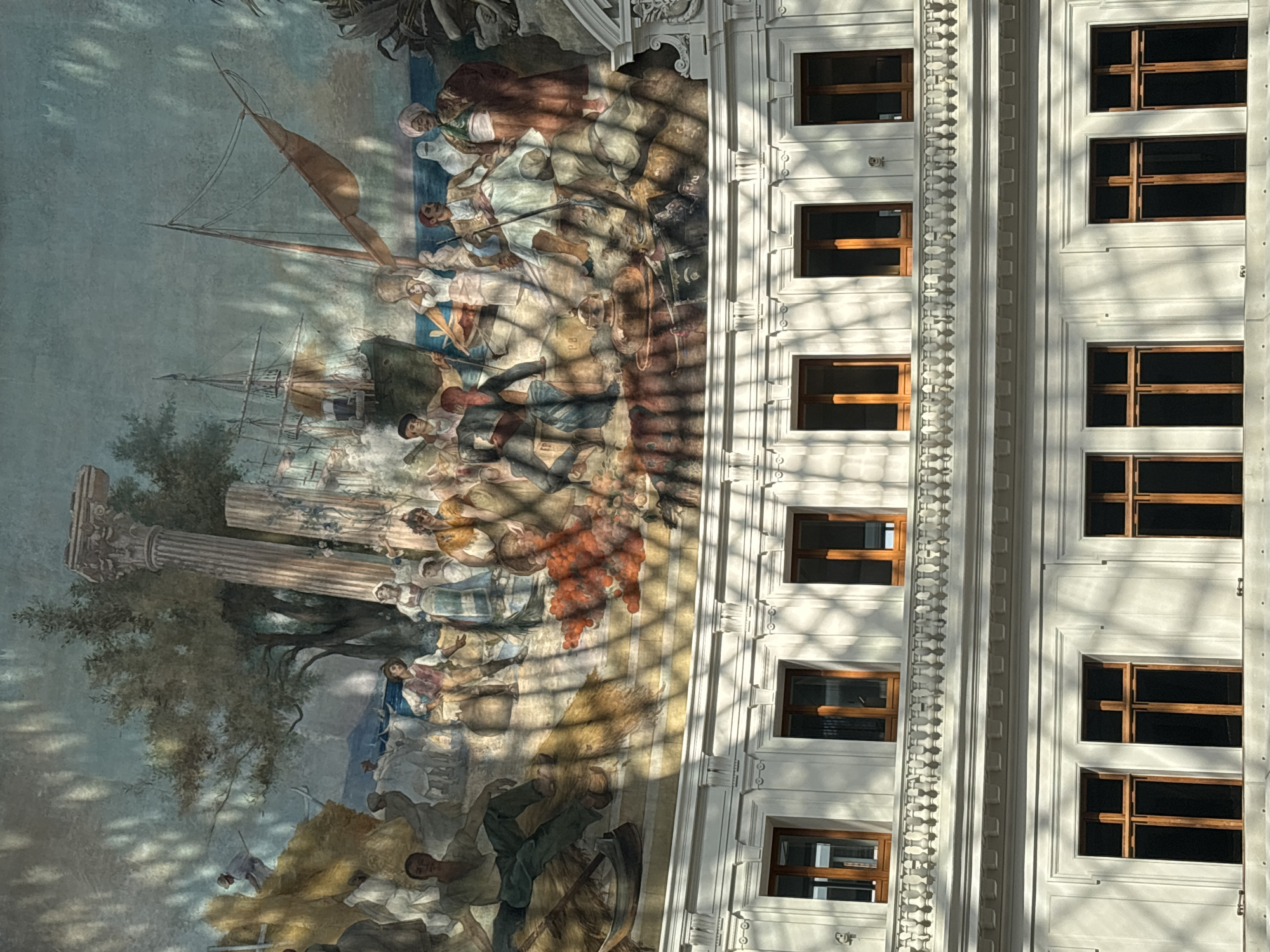 The image shows a grand fresco adorning the interior of a building in Paris, France. The fresco depicts a bustling scene of people engaged in various activities, with ships in the background, suggesting a maritime or historical theme. The artwork is illuminated by sunlight filtering through a latticed ceiling, casting a patterned shadow across the mural and the architectural elements below. The lower portion of the image reveals a facade with multiple windows, characteristic of Parisian architecture, with ornate balconies and decorative mouldings. The overall impression is one of historical grandeur and architectural beauty.