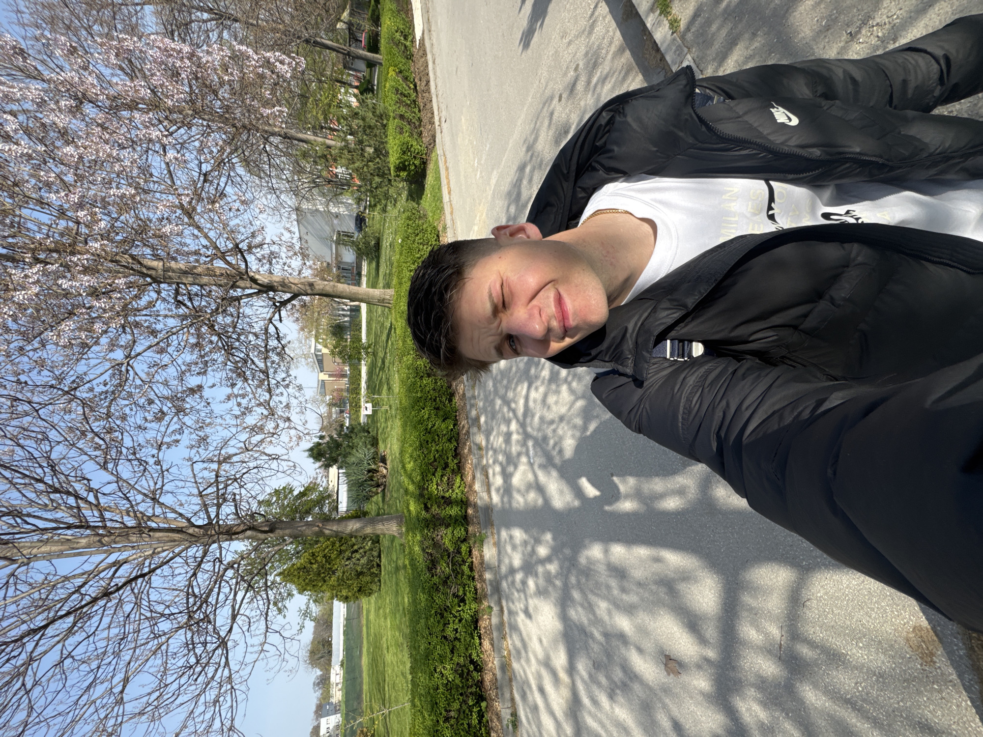 A selfie shows a young man with short dark hair, wearing a white T-shirt under a black jacket. He is squinting slightly with a smile. He appears to be standing on a sidewalk in a park or residential area in the city of Plovdiv, Bulgaria. The background shows trees, a grassy area, some buildings, and a blue sky. The lighting suggests it is daytime. The sidewalk he is standing on is beside a green hedge.