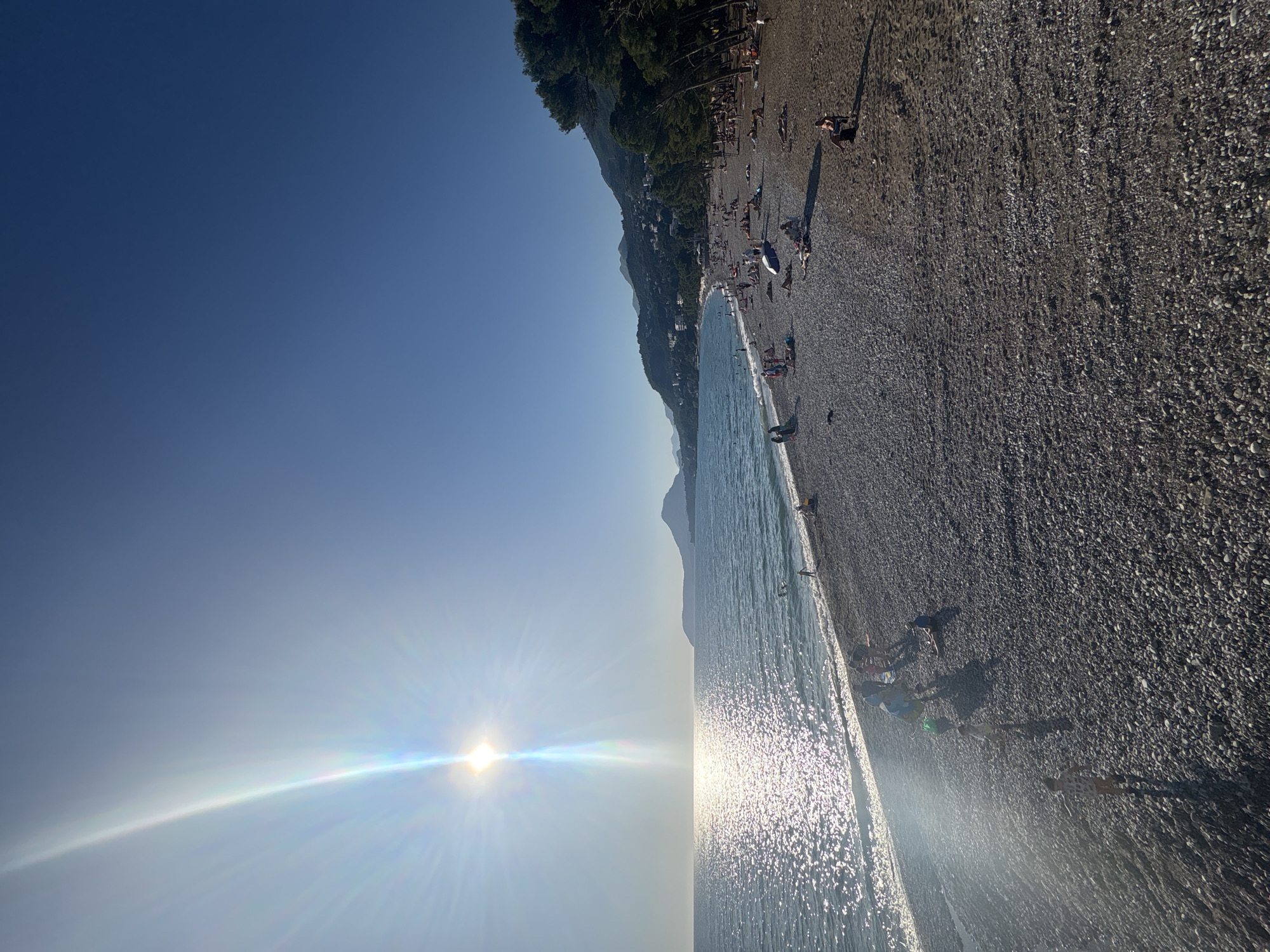 The image depicts a wide-angle view of a pebble beach in Bar, Montenegro, under a clear, bright blue sky with a low sun positioned over the sea on the left, generating a prominent lens flare and sunbeams. The sparkling glint of the sun is intensely reflected on the ocean's surface.
The beach itself is composed of dark grey pebbles, extending across the lower half of the frame. Numerous individuals are scattered across the beach. On the left side, near the waterline, a group of people, including several children, are standing or wading. One small child, wearing patterned shorts and a shirt, stands alone on the pebbles, facing away from the camera, casting a long shadow to the right.
Further along the beach to the right, many people are sunbathing, lying on towels or directly on the pebbles. A white sun umbrella is open towards the center of the beach, with individuals lounging nearby. Towards the far right, a line of tall, dark green coniferous trees borders the beach, casting long shadows onto the sand where more people are relaxing, some under the shade. One person is seated in a dark beach chair.
In the sea, several individuals are visible, either swimming or wading near the shore.
The background features a verdant hillside densely covered with vegetation and numerous white-colored buildings, indicating a coastal town or settlement. Beyond the hillside, a range of mountains stretches into the distance, with one prominent, somewhat conical mountain visible across the bay on the left horizon. The lighting suggests late afternoon, with the sun low in the sky and casting extended shadows.