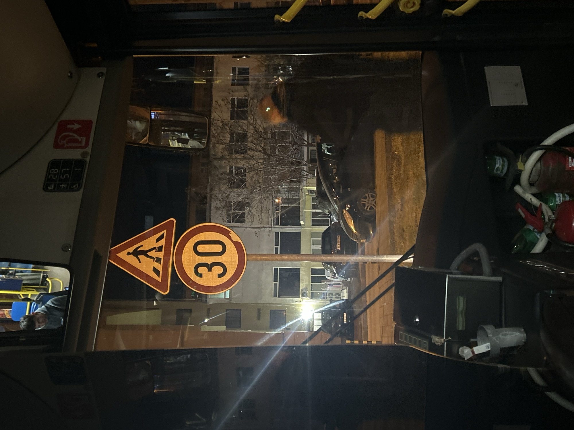 The image is taken from inside a bus at night in Varna, Bulgaria. The windshield shows street signs for a pedestrian crossing and a 30 km/h speed limit. Outside, a building with multiple windows and a bare tree branch are visible. Cars are parked along the street. A rear-view mirror reflects the bus interior. Inside the bus, there's a fire extinguisher and various equipment next to the driver's seat. A passenger is visible in another window reflection.