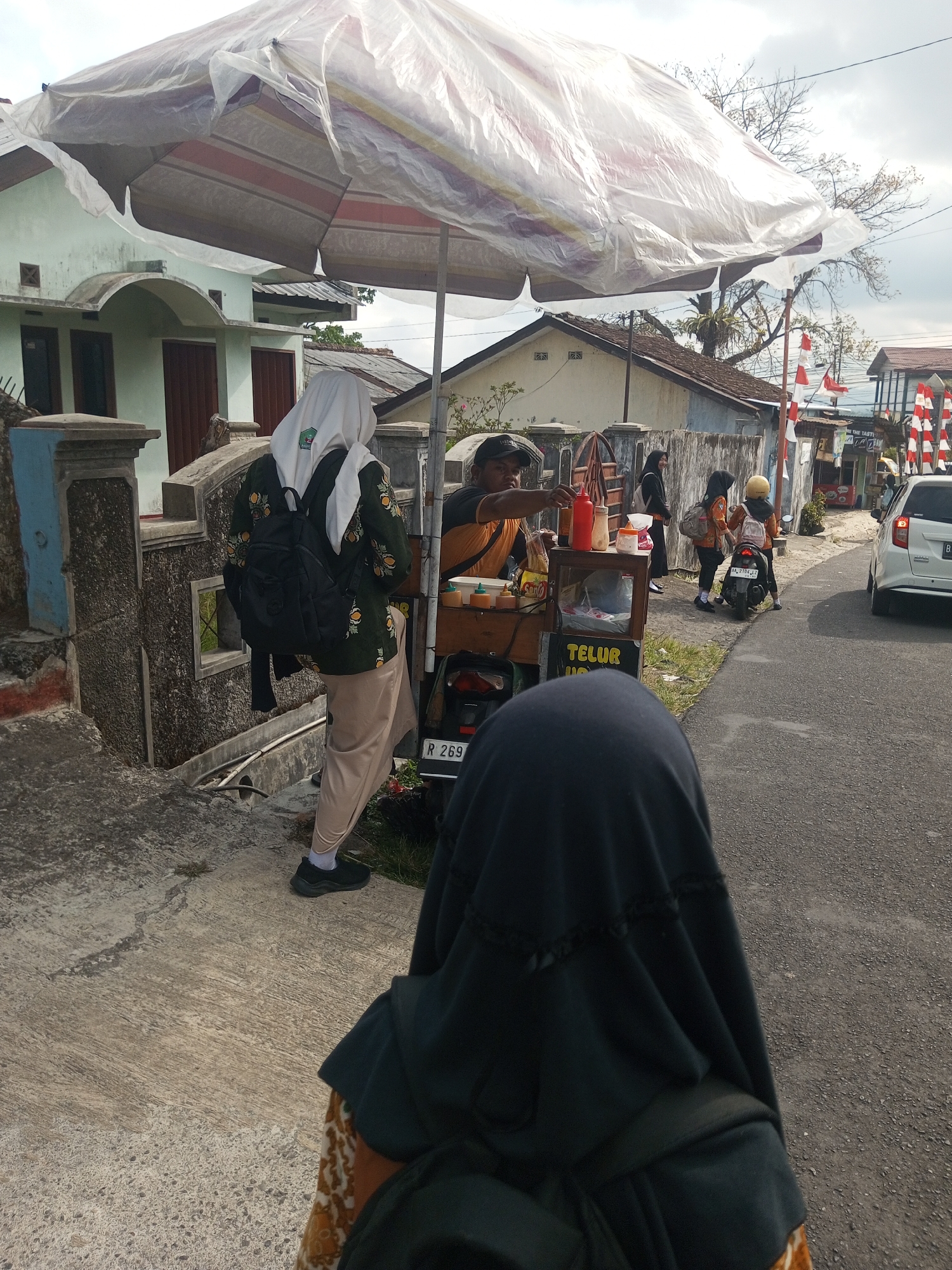 In Krasak, Indonesia, a street vendor sells food from a cart under a large, makeshift umbrella. The vendor, a man wearing an orange shirt and a black cap, is actively serving customers. Two young women, dressed in hijabs and modest clothing, are interacting with the vendor and the food cart. One woman, wearing a white hijab and a green jacket with floral patterns, stands with her back to the camera, a black backpack slung over her shoulders. The other woman, closer to the food cart and facing away from the viewer, wears a black hijab and appears to be a customer. A motorcycle is parked behind the women, and a white car is visible on the road in the background. The environment suggests a typical urban street scene, with houses and a road lined with Indonesian flags. The weather appears to be overcast.