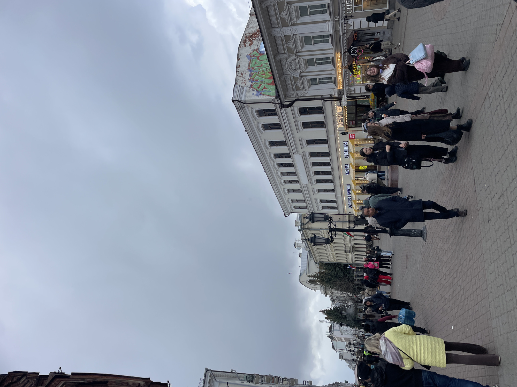 This image captures a vibrant street scene in Kazan, Russia, under an overcast sky. The street is a pedestrian zone, paved with light-colored bricks or cobblestones, and is lined with multi-story buildings exhibiting classic architectural styles, including ornate facades and numerous windows.
Several commercial establishments are visible, with signs featuring Cyrillic text. Notable signs include "HOTEL JOY", "ТЫРЛОТ", "СУВЕНИРЫ" (Souvenirs), and "ПАТШАК". Partial text like "ЧЕХ" and "ЗДРАВСТВУЙТЕ" (Hello) can also be seen. A prominent detail on the upper part of a building to the right is a large section of green and red graffiti. Black, ornate lampposts are spaced along the street.
Numerous people are seen walking in various directions, indicating a busy urban environment. They are dressed in coats, jackets, and long sleeves, suggesting cool or cold weather. Individuals carry bags, including a bright pink backpack on one person in the mid-ground and a person in the foreground wearing a distinctive bright yellow quilted coat. The atmosphere appears to be one of everyday activity, with people going about their routines. The cloudy sky suggests a daytime setting, though specific time is indeterminate due to the lack of direct sunlight or strong shadows.