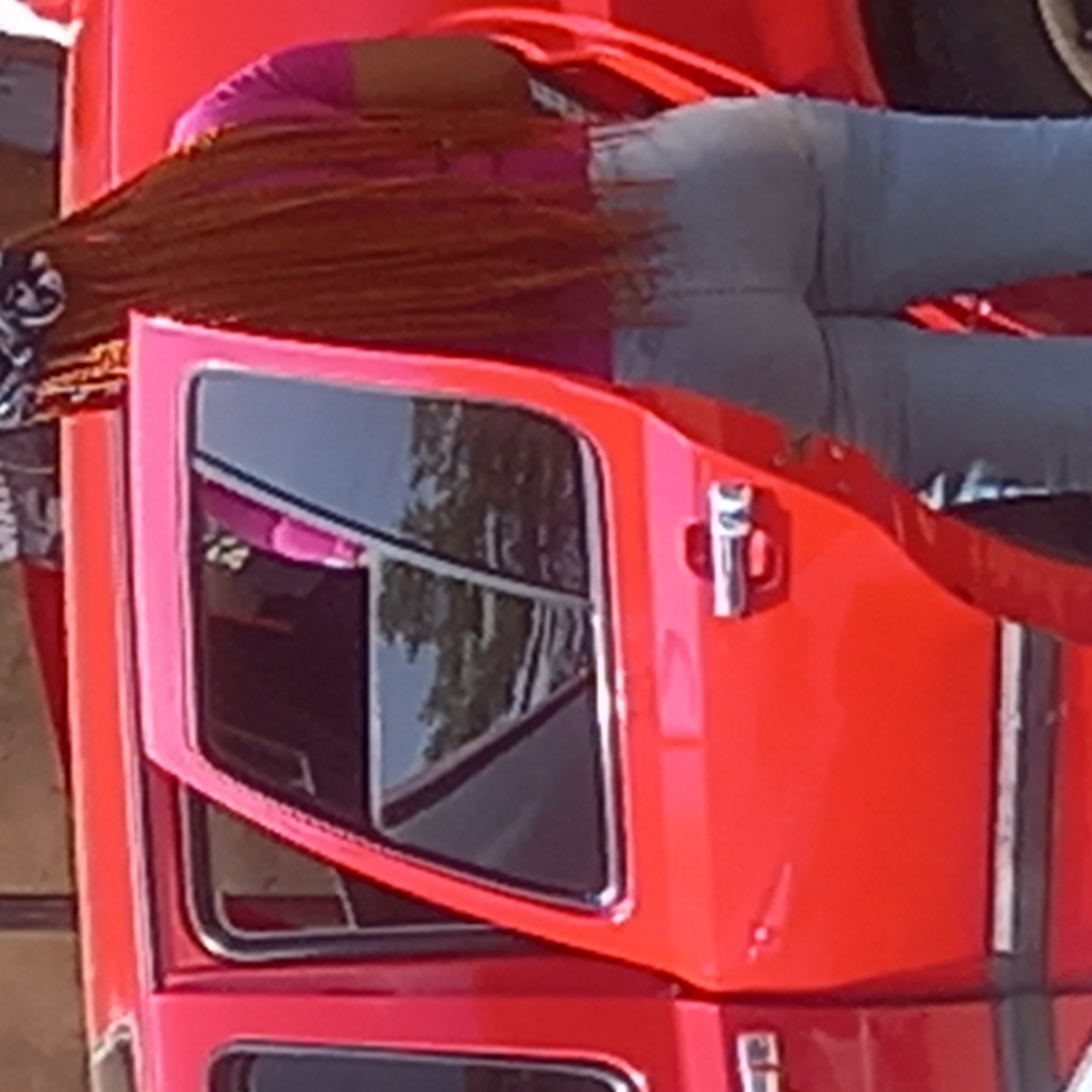 The image captures an outdoor scene featuring a person interacting with a vibrant red truck, set in Dadi Riba, Nigeria. The photograph, presented rotated, shows the passenger side of a large, red vehicle, likely a truck or utility van, with a person leaning into its open cab door.
The person, possibly female, is dressed in a bright pink top and light blue or grey jeans. Her upper body is bent over the door frame, with her head obscured. Long, distinctive reddish-brown braided hair falls prominently from her head towards the vehicle's interior. A silver door handle is visible on the open door.
The red vehicle is a strong focal point, featuring bright pink trim around its window frames, adding a colorful contrast. The dark tinted windows reflect the surrounding environment, showing clear outlines of trees and foliage, suggesting a sunny daytime setting with good weather. The person appears to be either entering the vehicle, exiting, or reaching for something inside. No specific emotions are discernible, as the person's face is not visible. There is no legible text visible on the vehicle or the person's clothing.