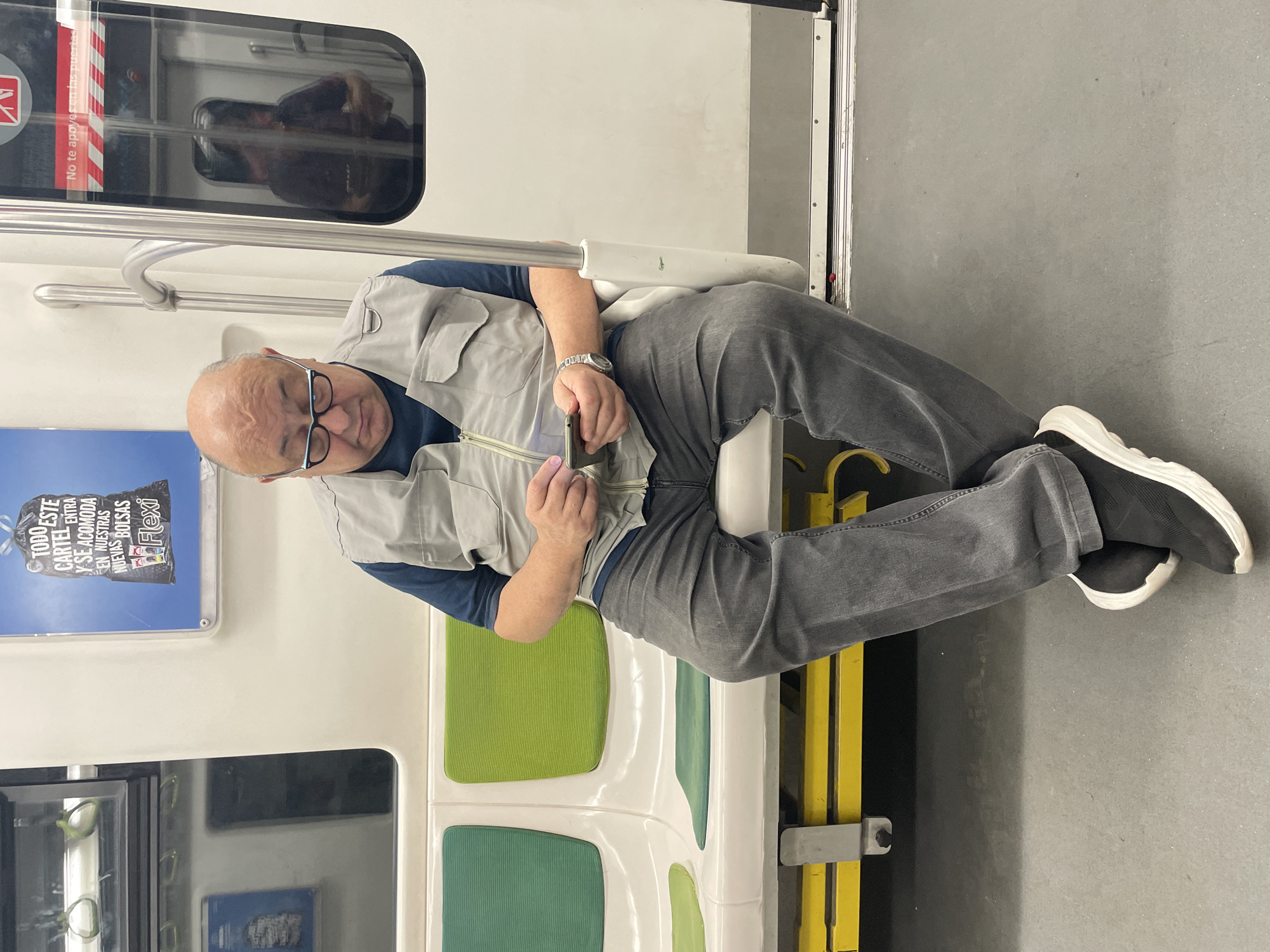 An older man sits on a green and white train seat, engrossed in his phone. He wears glasses, a gray vest over a navy shirt, gray jeans, and black and white sneakers. The interior of the train is predominantly white, with a sign visible above him written in Spanish. The doors and windows are to his left, and a gray floor is to his right. The window has a sticker on it warning against leaning on the doors. The sign shows an advertisement for Flexi shoes. The setting is likely a subway train car in Buenos Aires, Argentina, given the Spanish signage.