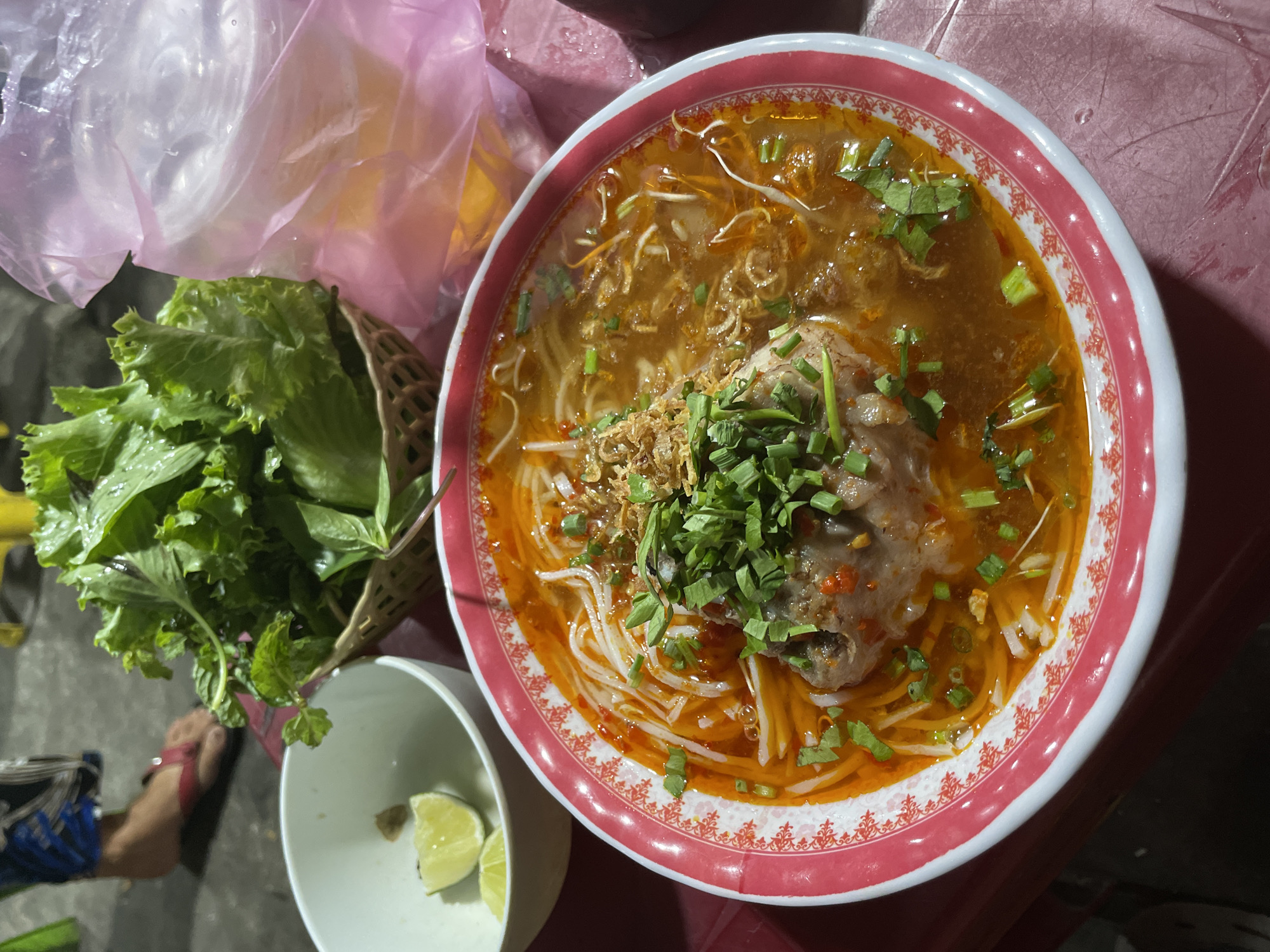 The image shows a bowl of Vietnamese noodle soup, likely Bun Bo Hue, on a red table. The soup contains white noodles, broth, meat, and green herbs. Next to the soup are a basket of lettuce and basil leaves, and a small bowl with lime wedges. A plastic bag sits behind the basket of lettuce. The setting appears to be outdoors, possibly a street-side food stall. A person's leg and foot are visible in the lower left corner. The location is Binh Thuan Ward, Vietnam.
