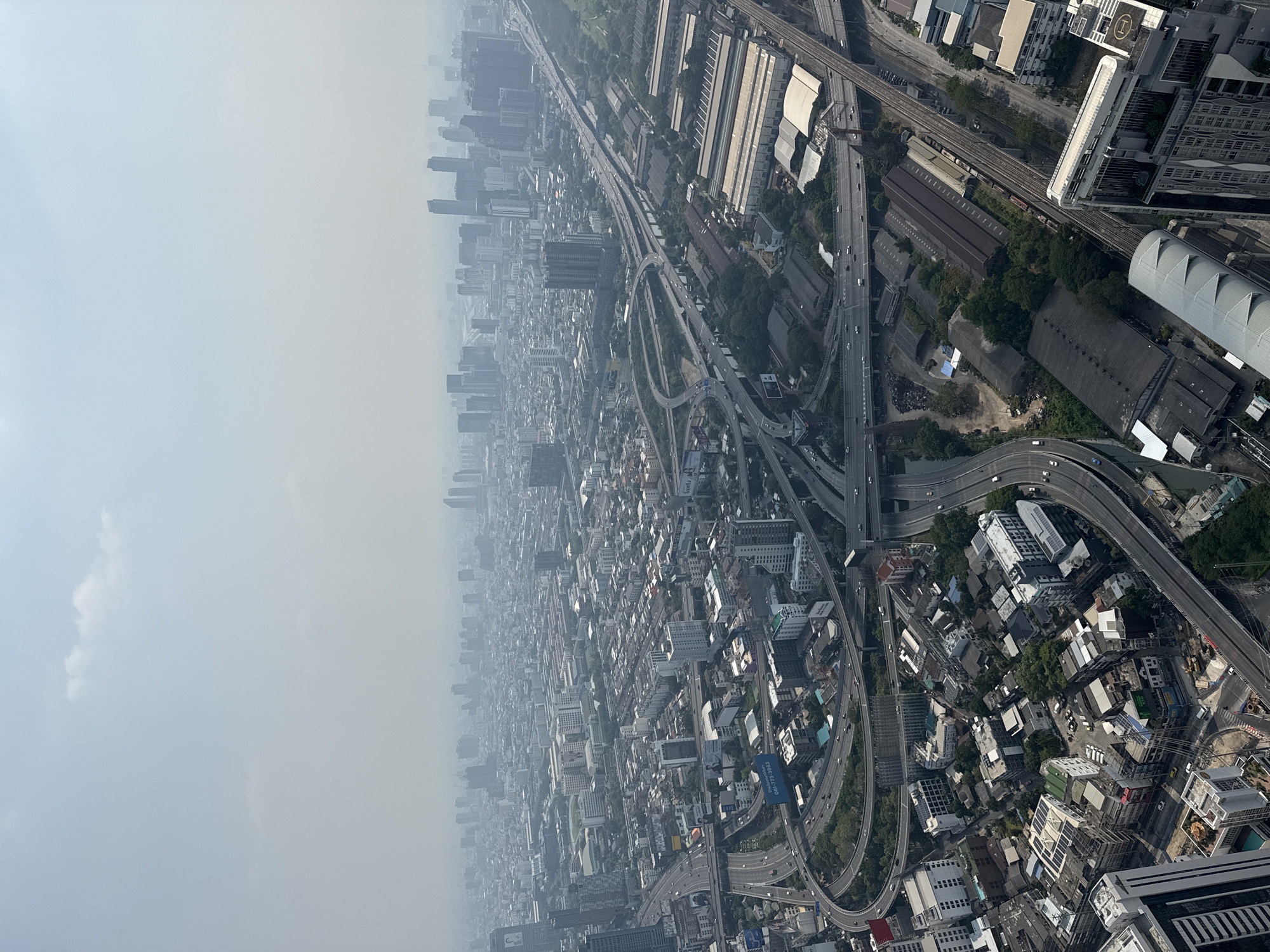 The image shows an aerial view of Bangkok, Thailand. The city is densely populated with a mix of low-rise and high-rise buildings. Many roads and highways weave through the city, some elevated. The air is hazy or polluted, reducing visibility. No people are visible. The scene depicts urban infrastructure and environmental conditions in Bangkok.