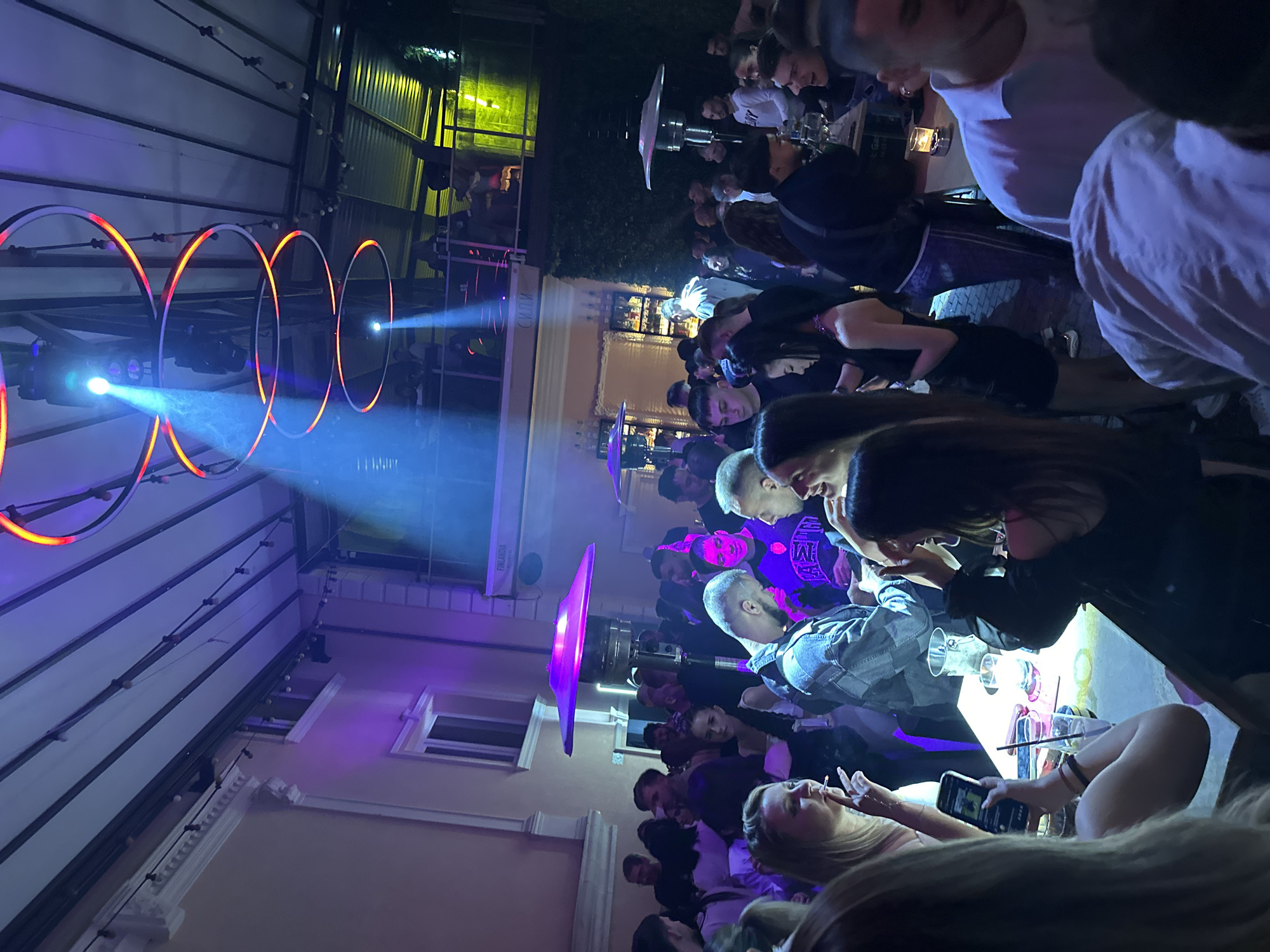 A nighttime scene shows a crowded outdoor area, likely a restaurant or bar, in Plovdiv, Bulgaria. Multiple groups of people are seated at tables. Lighting is provided by overhead fixtures, including circular neon lights and spotlights. Some individuals are smoking, while others are using their phones. The environment includes building exteriors and covered overhead structures. The overall atmosphere is lively and social.
