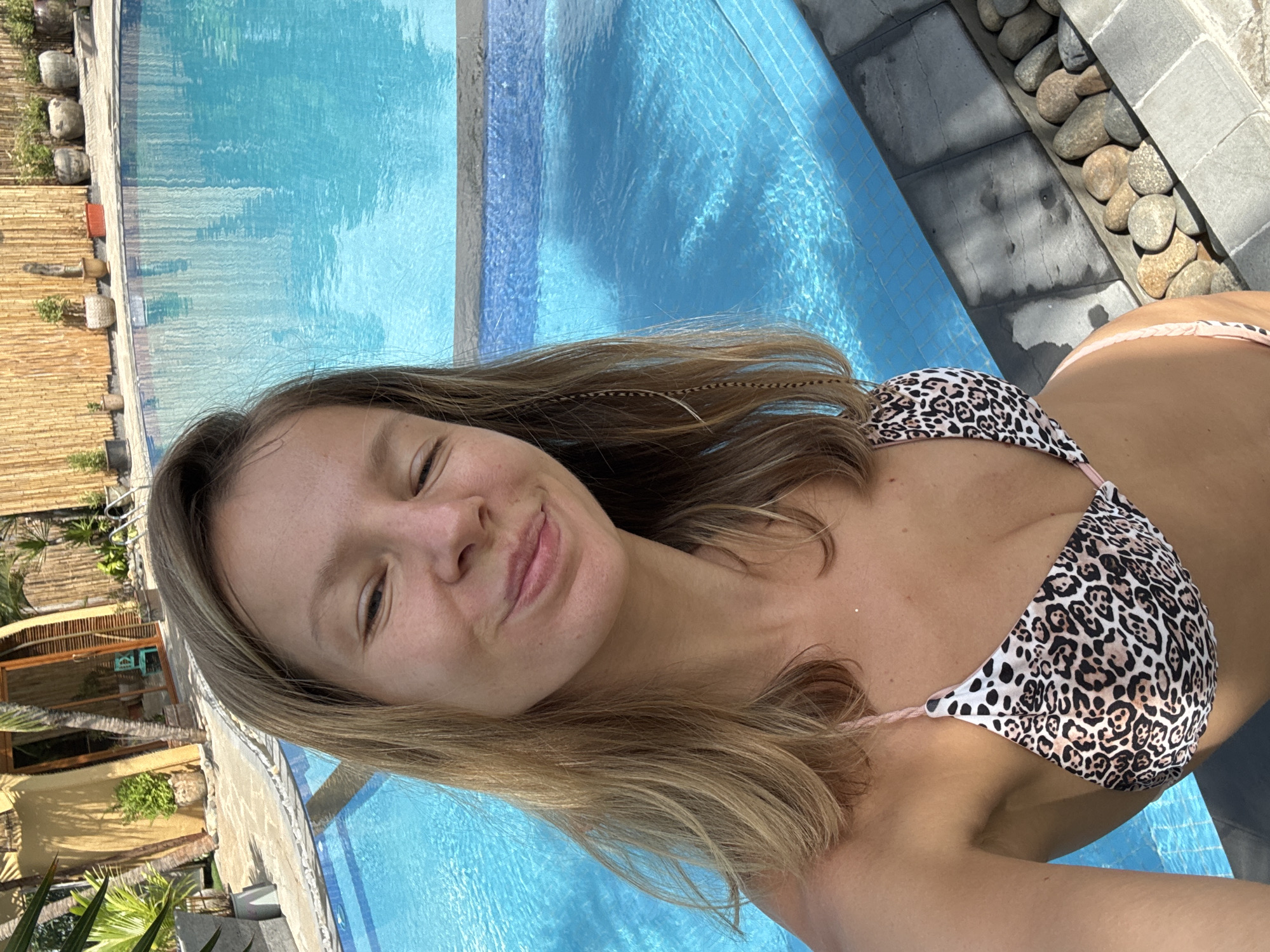 A woman in a leopard print bikini is taking a selfie by a swimming pool. She has blonde hair and is smiling. The pool is blue and surrounded by a stone walkway and lush greenery. In the background, there is a building with a thatched roof and bamboo accents, typical of the architecture found in Mũi Né, Vietnam. The overall scene suggests a relaxed vacation atmosphere.