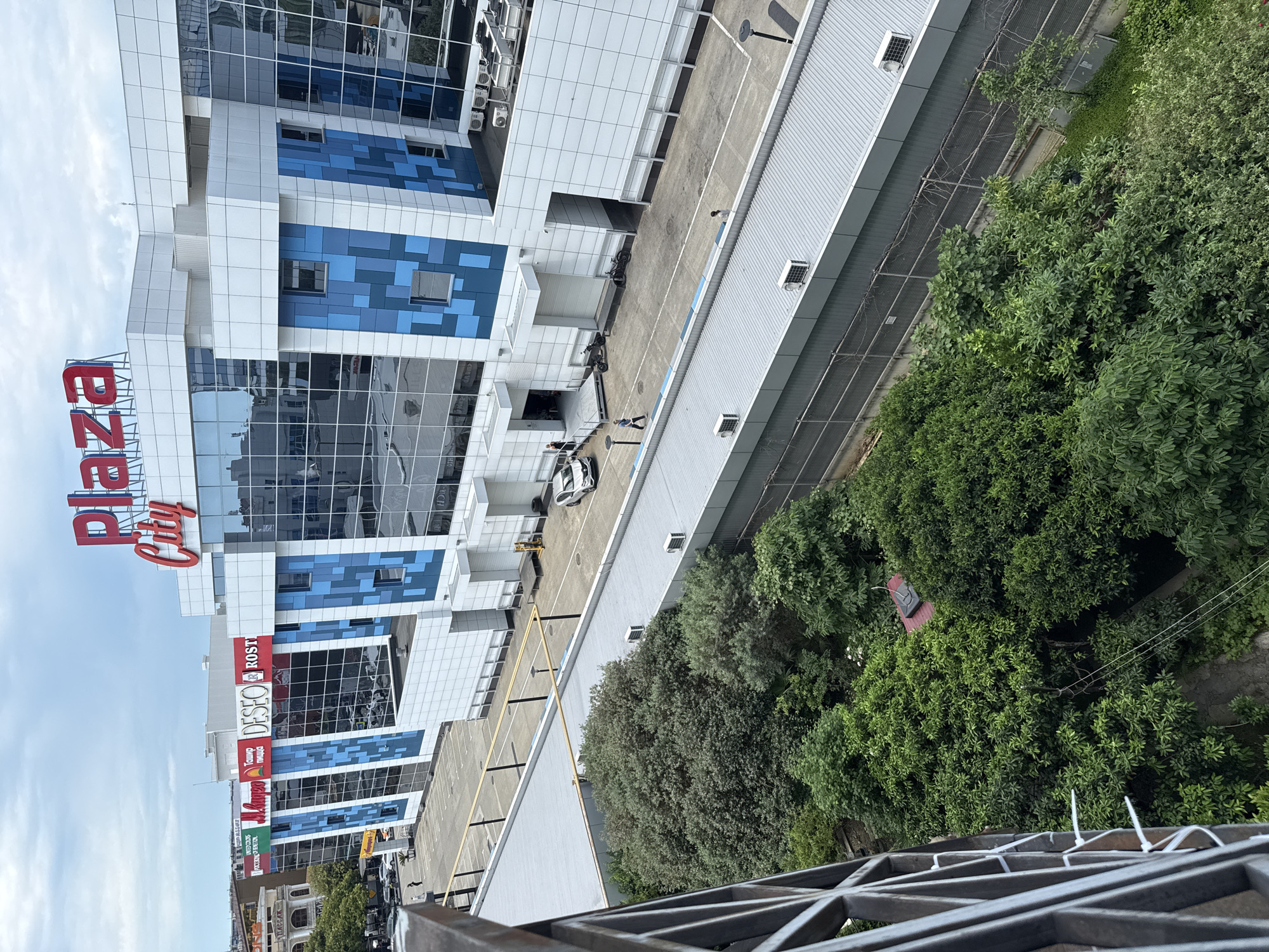 The image depicts the Plaza City shopping center in Sochi, Russia, on a cloudy day. The facade of the modern building features large glass windows with blue geometric accents. Above the entrance, the red lettering "Plaza City" is prominently displayed. To the left of the main building, a row of shops with colorful signs, including "Mango" and "DESEO," are visible. In the foreground, lush green trees and foliage are abundant, suggesting a well-maintained urban environment. A paved area in front of the shopping center shows parked vehicles and a few individuals. The scene is captured from an elevated viewpoint, possibly from an adjacent building or balcony, with a metal railing visible in the lower left corner.