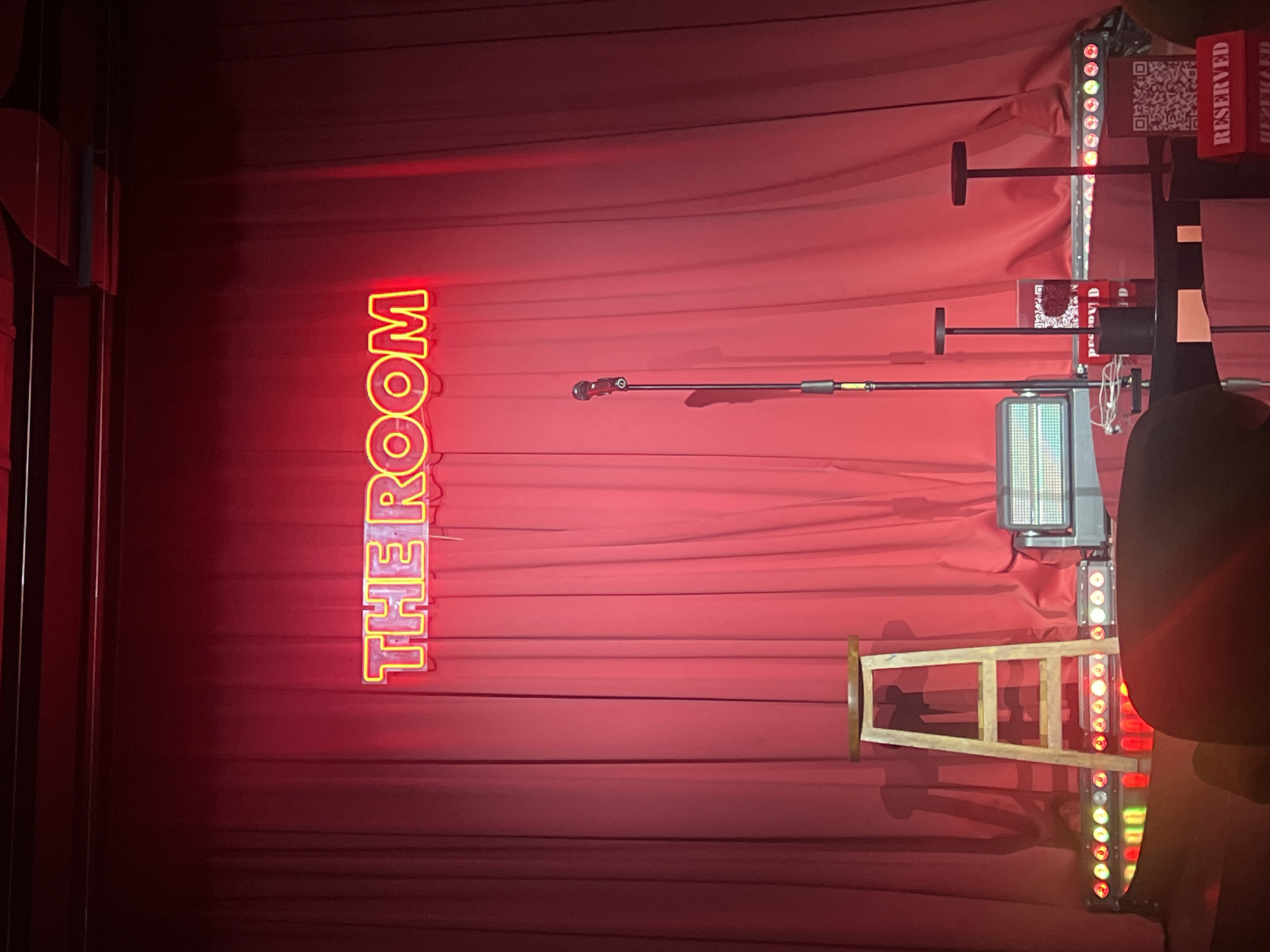 The image depicts a dimly lit indoor space, possibly a performance venue, stage, or club, with a dominant red curtain backdrop. The scene is rotated 90 degrees counter-clockwise.
A prominent vertical neon sign on the left side of the red curtain spells out "THE ROOM" in bright red letters with a yellow outline. Across the middle of the frame, a black microphone stand extends horizontally. Below it, a metallic, rectangular fixture, possibly a stage light or projector with a grid-like surface, is visible.
On the right side of the frame, multiple vertical strips of LED lights are mounted, displaying various colors including red, green, and white. Several red rectangular signs, each bearing the word "RESERVED" in white text and a QR code, are attached to poles and positioned towards the right. A light-colored wooden stool or chair leans against the curtain near the bottom right corner. A dark, indistinct form, possibly part of a person or equipment, is partially visible on the far right.
The overall setting suggests a space designed for live performances or events, characterized by its stage lighting, microphone, and a backdrop that creates a theatrical atmosphere. The "RESERVED" signs indicate designated seating or VIP areas. The scene is likely located in North Kuta, Indonesia, and takes place in the evening or a darkened environment.