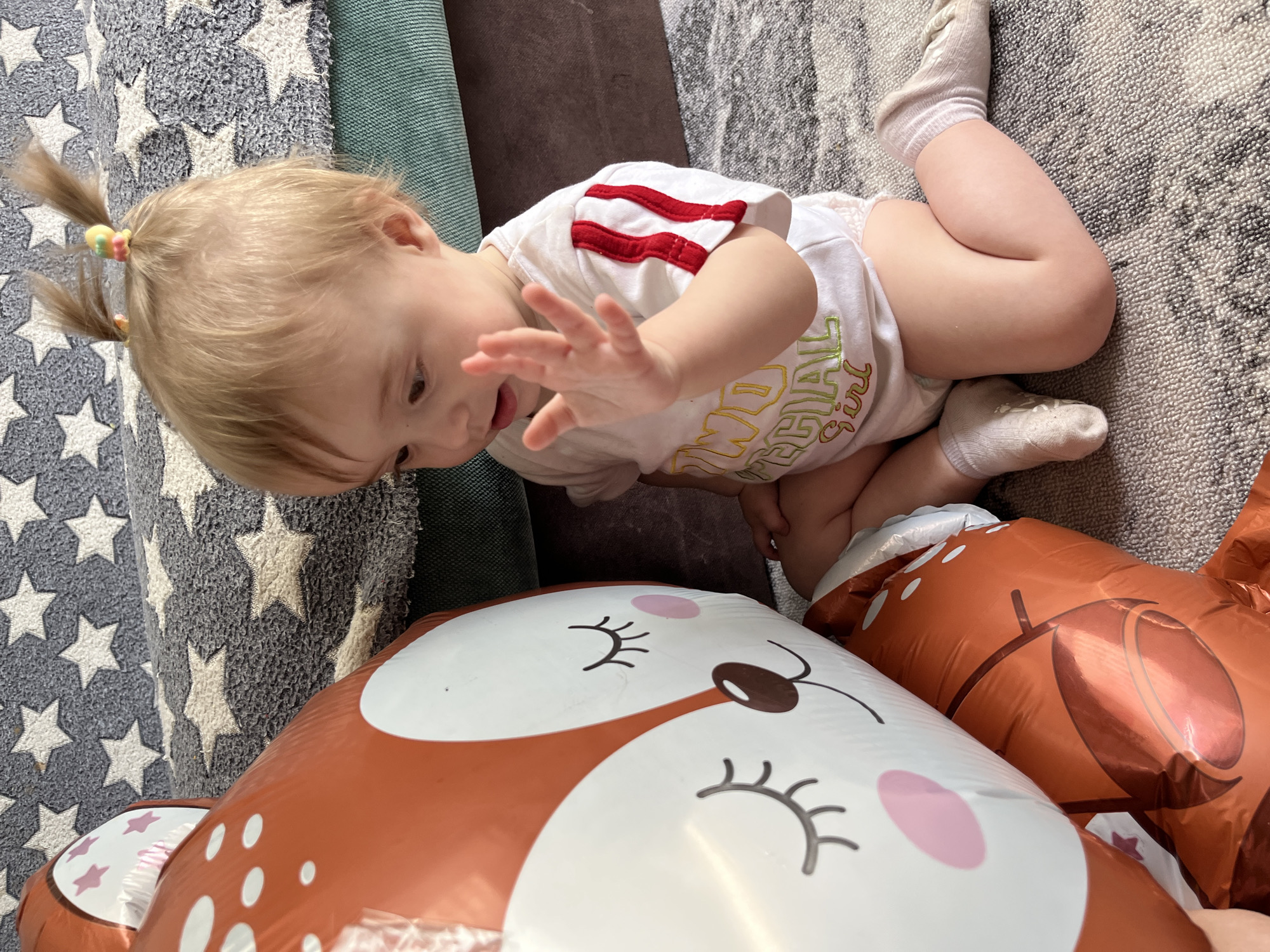 A young child with blonde hair styled in two ponytails, each secured with a colorful hair tie, is kneeling on a carpet with a pattern of gray and white stars alongside a gray carpet, appearing to reach towards a large, inflatable foil balloon shaped like a fox. The child is wearing a white onesie with red stripes on the sleeves and a colorful design on the front, as well as white socks. The environment suggests an indoor setting. No information is available about Chaikovsky, Russia.