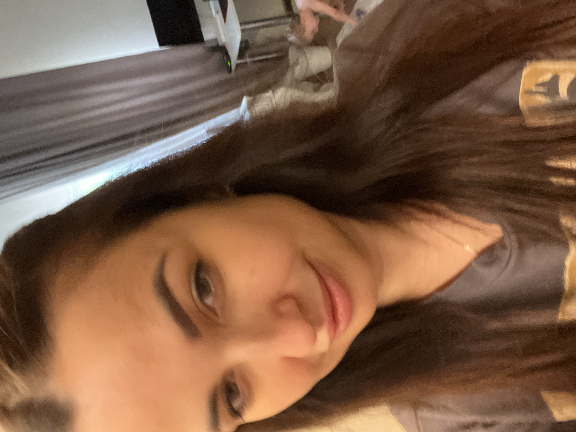 A woman with long, dark brown hair is taking a selfie. She is wearing a dark grey t-shirt with a gold graphic on it and a delicate gold necklace. Her hair is down, and she is looking towards the camera with a slight smile. The background is slightly out of focus and shows a curtain, a white trash can, a desk with a computer monitor, and a small child playing on the floor. The lighting is warm and appears to be indoors.