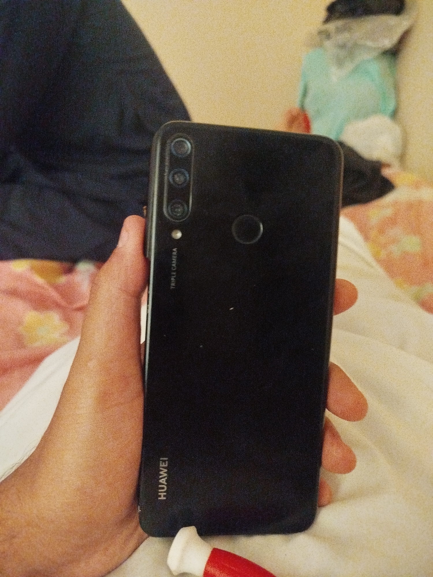 A hand holds a black smartphone with "HUAWEI" text at the bottom and "TRIPLE CAMERA" text near the top. The phone's rear features a triple camera setup and a circular fingerprint sensor. The background is blurred, showing dark fabric, light-colored bedding with a floral pattern, and a light-colored wall. The scene appears to be indoors, possibly in a bedroom. There is no indication of location in Lassan, Pakistan.
