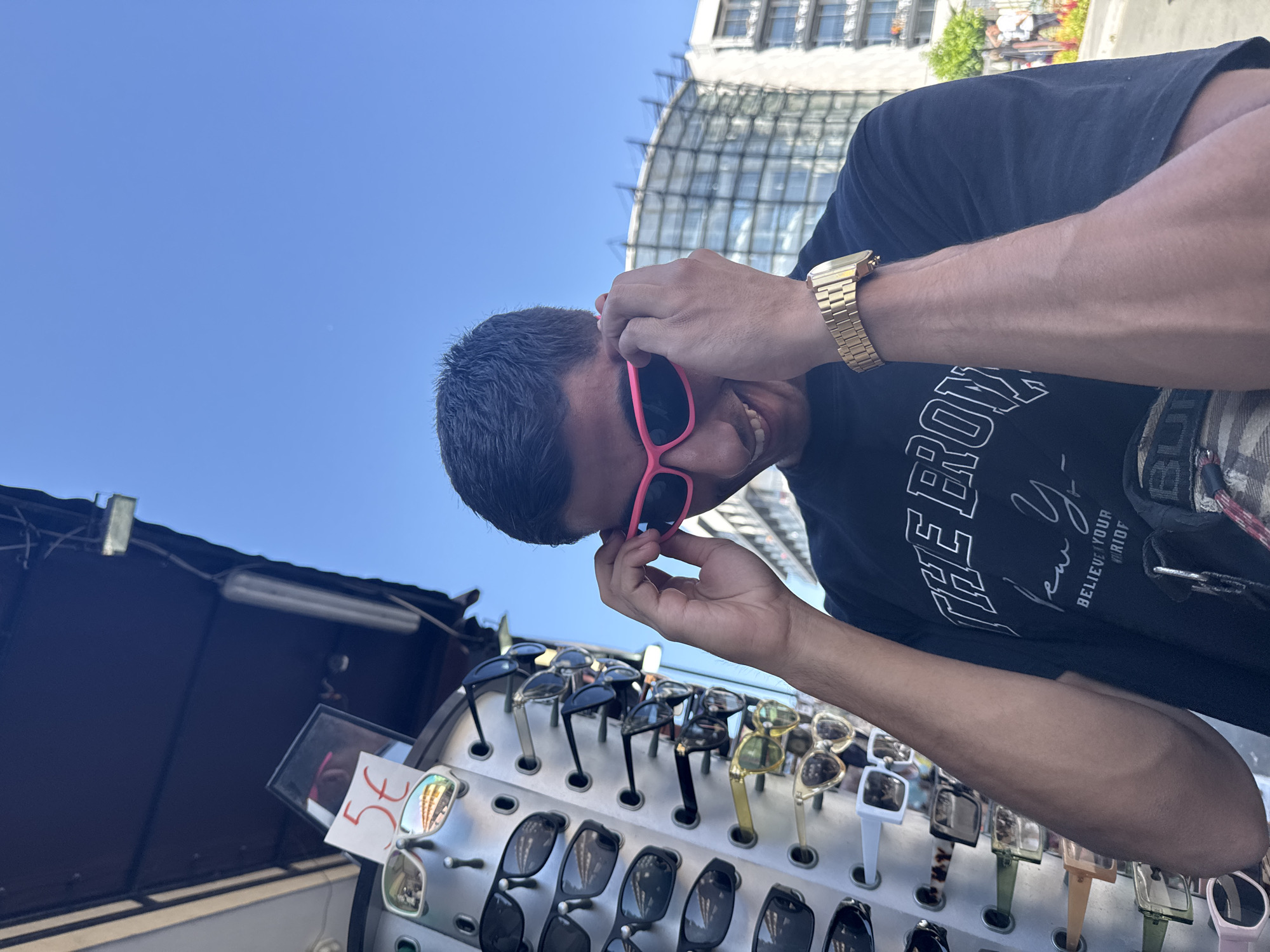 A young man wearing a black t-shirt and a gold watch is trying on pink sunglasses in front of a display of sunglasses for sale. The display rack is full of various styles of sunglasses. In the background, a modern building with a glass facade and blue sky can be seen. The scene appears to be outdoors, possibly on a street or market in Nesebar, Bulgaria, as indicated by the price tag on the display.