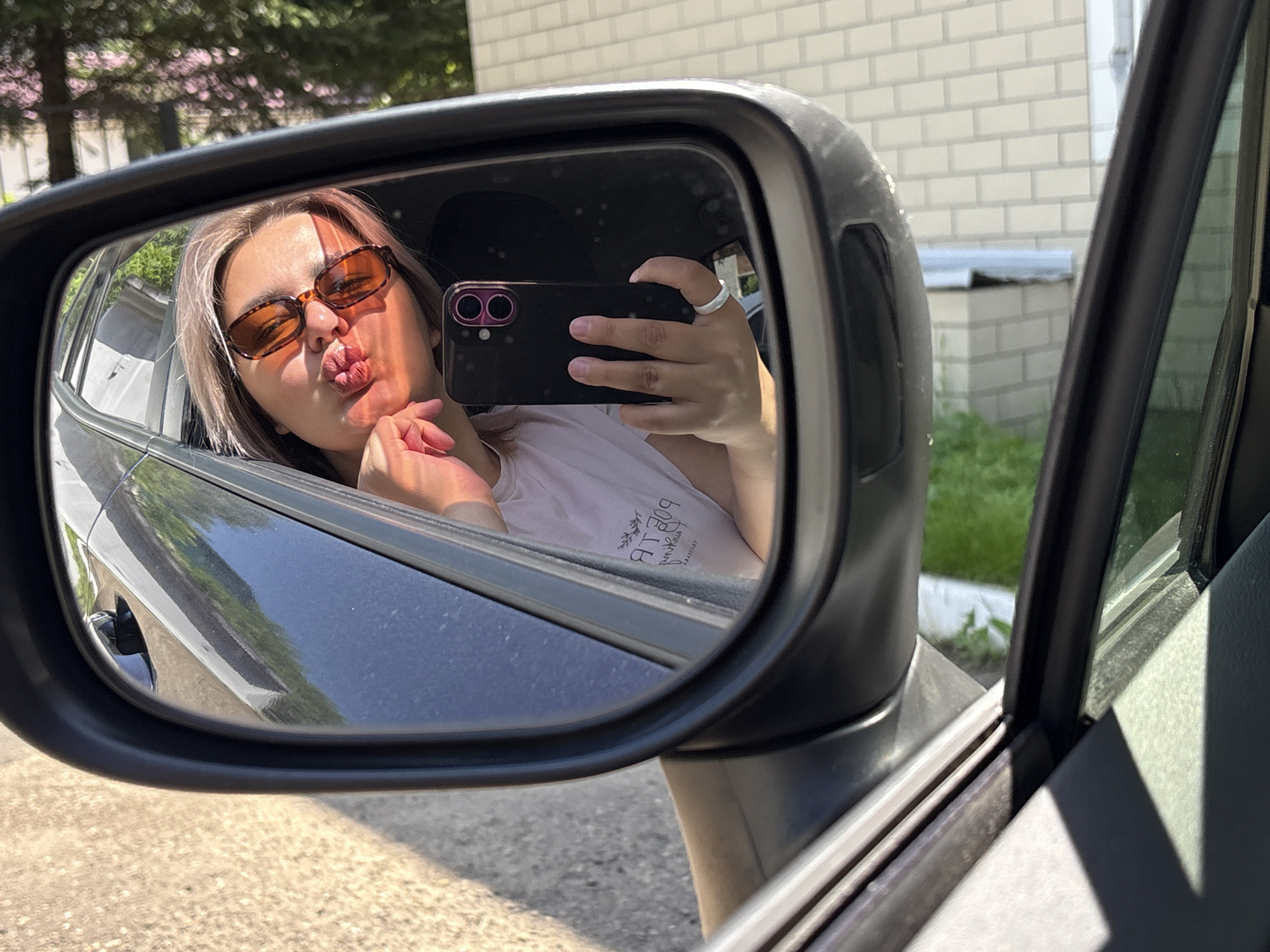 A young woman with blonde hair and sunglasses is reflected in a car's side mirror. She is leaning out of the car window, holding a smartphone to take a selfie, and making a kissing face. Her hair is styled with bangs, and she is wearing a white t-shirt. The reflection also shows the interior of the car and the exterior environment. Outside the car, a brick building and green foliage are visible. The scene is illuminated by daylight, casting shadows. The photograph appears to have been taken on a sunny day, possibly in the city of Birobidzhan, Russia, given the information provided.