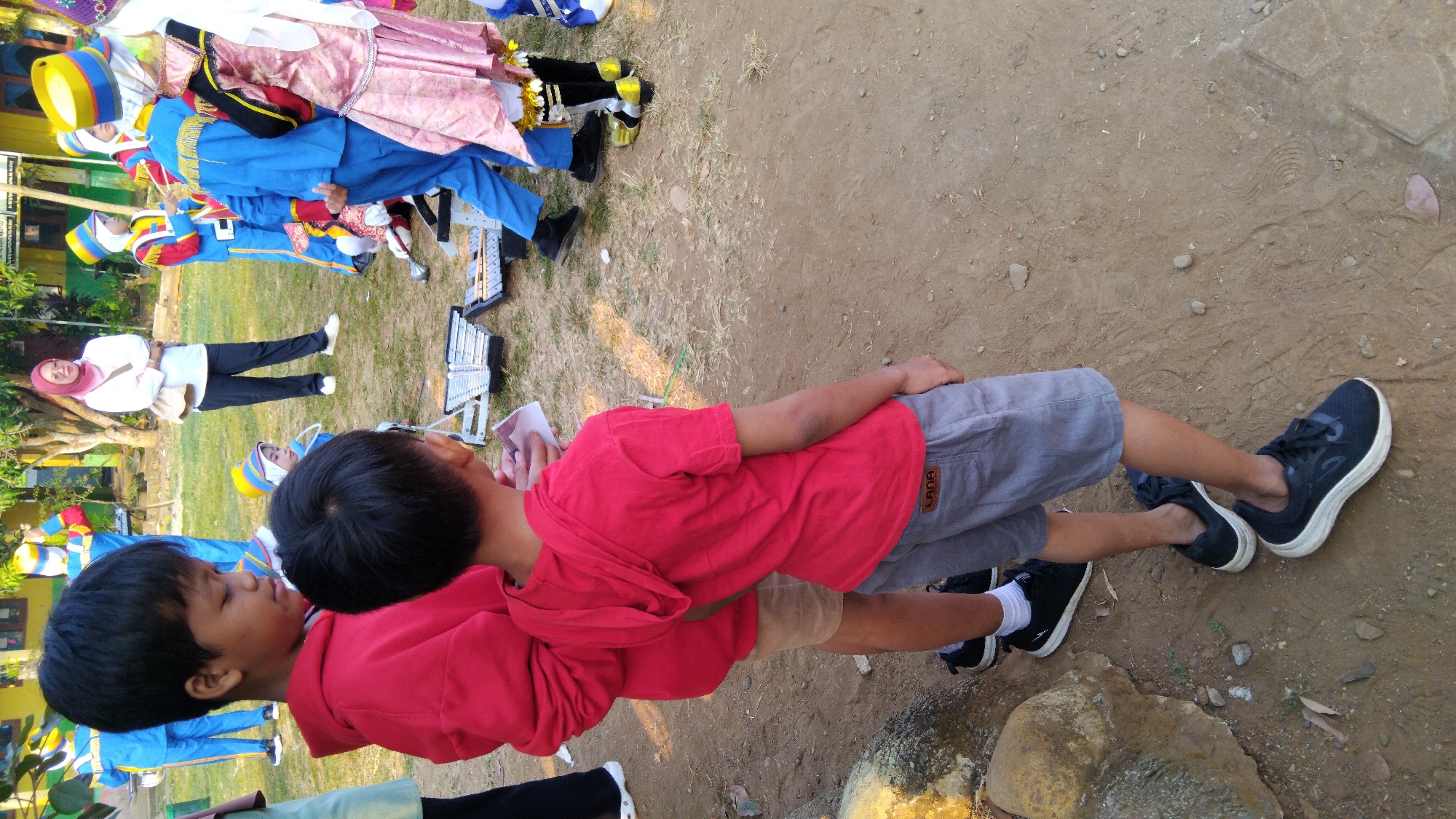 In Sembojo, Indonesia, on a dirt field with dry grass and scattered rocks, two young boys stand facing away from the camera. The boy in the foreground wears a red hooded t-shirt and grey shorts, his dark hair is short. He is looking down at a piece of paper he holds in his hand. The boy behind him is wearing a red hooded sweatshirt. Other children in marching band uniforms are visible in the background, some wearing blue and yellow outfits, others in pink and gold. A woman in a pink headscarf and white shirt stands to the left, her arms crossed. A xylophone is visible on the ground between the children. The scene appears to be outdoors during daylight.