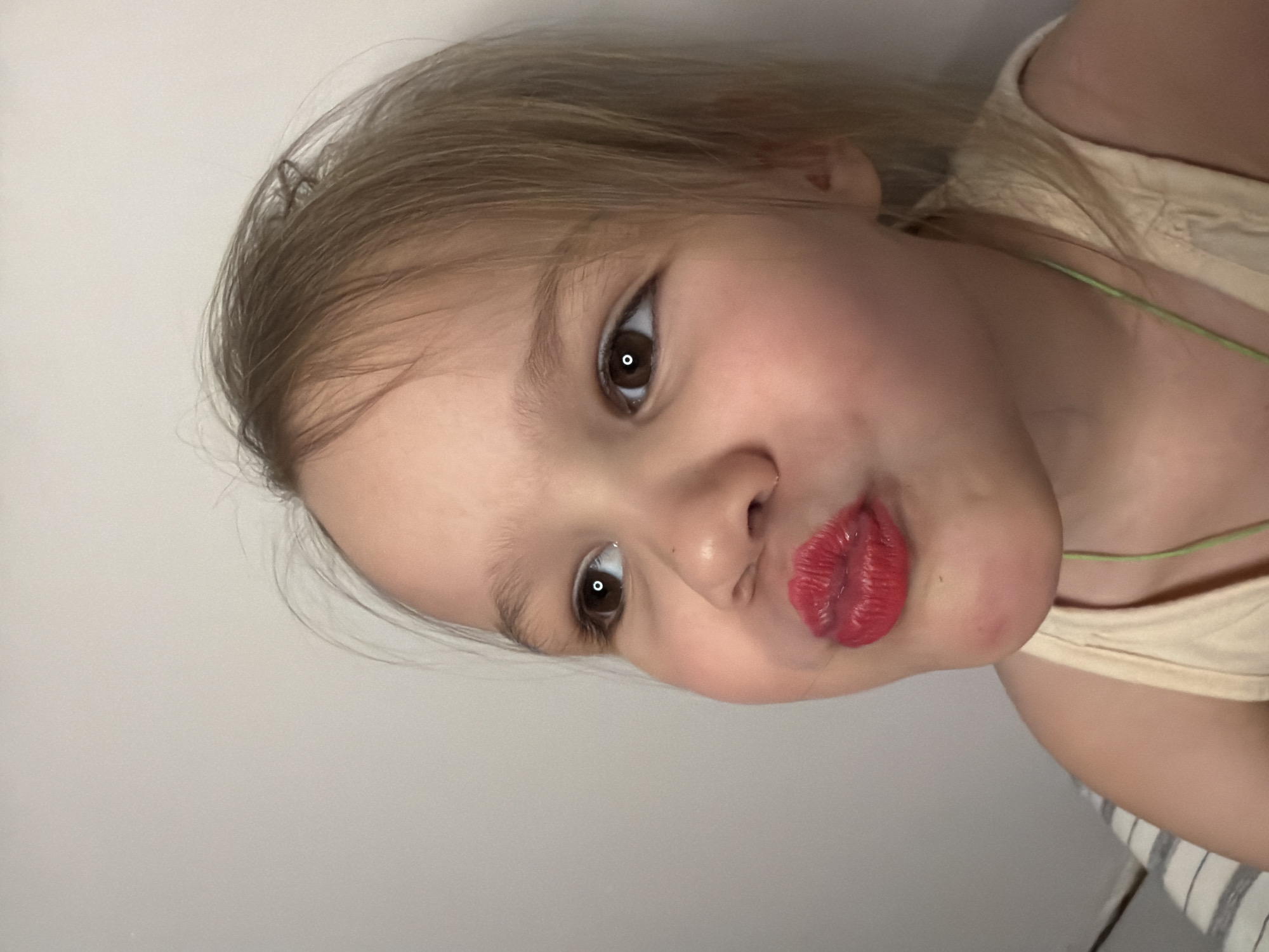 A close-up image shows a young child with light blonde hair, fair skin, and brown eyes. The child's face is tilted towards the camera, and they are pouting their lips, which are painted with bright red lipstick. The child is wearing a light beige or cream-colored top, and a thin green string is visible around their neck. The background is a plain white wall.