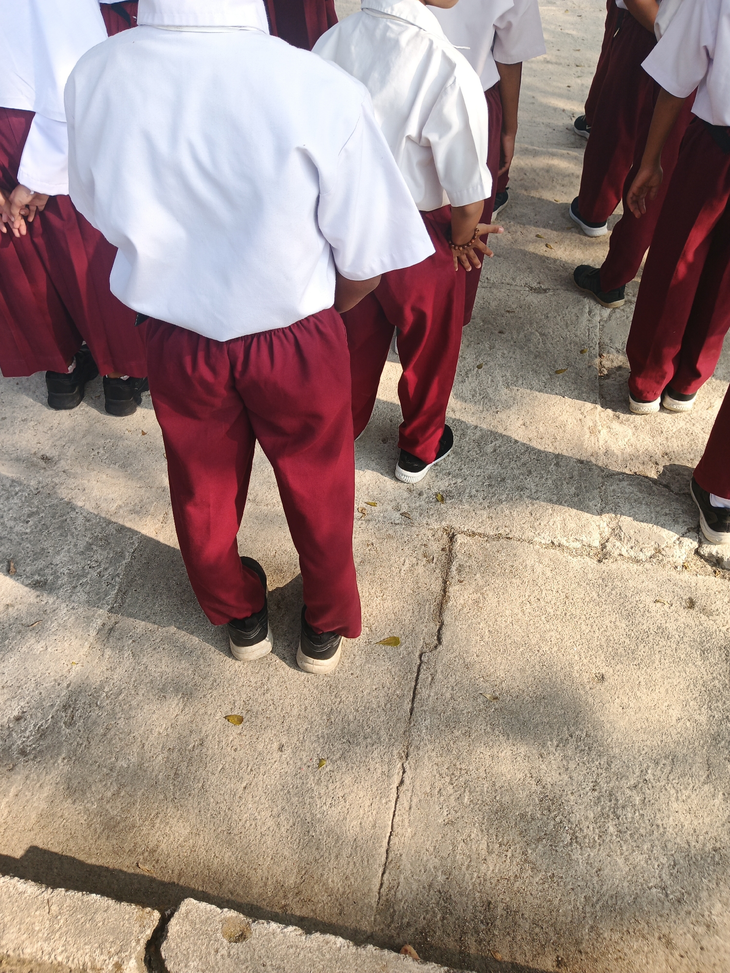 This image shows the lower bodies of several individuals standing on a concrete surface, likely outdoors. They are all wearing maroon-colored trousers and white shirts. The trousers appear to be school uniforms. The concrete surface has some visible cracks and is illuminated by sunlight, casting shadows. The setting is identified as the city of Rejo Basuki, Indonesia. The focus is on the legs and feet of the individuals, with some of them wearing black and white sneakers.