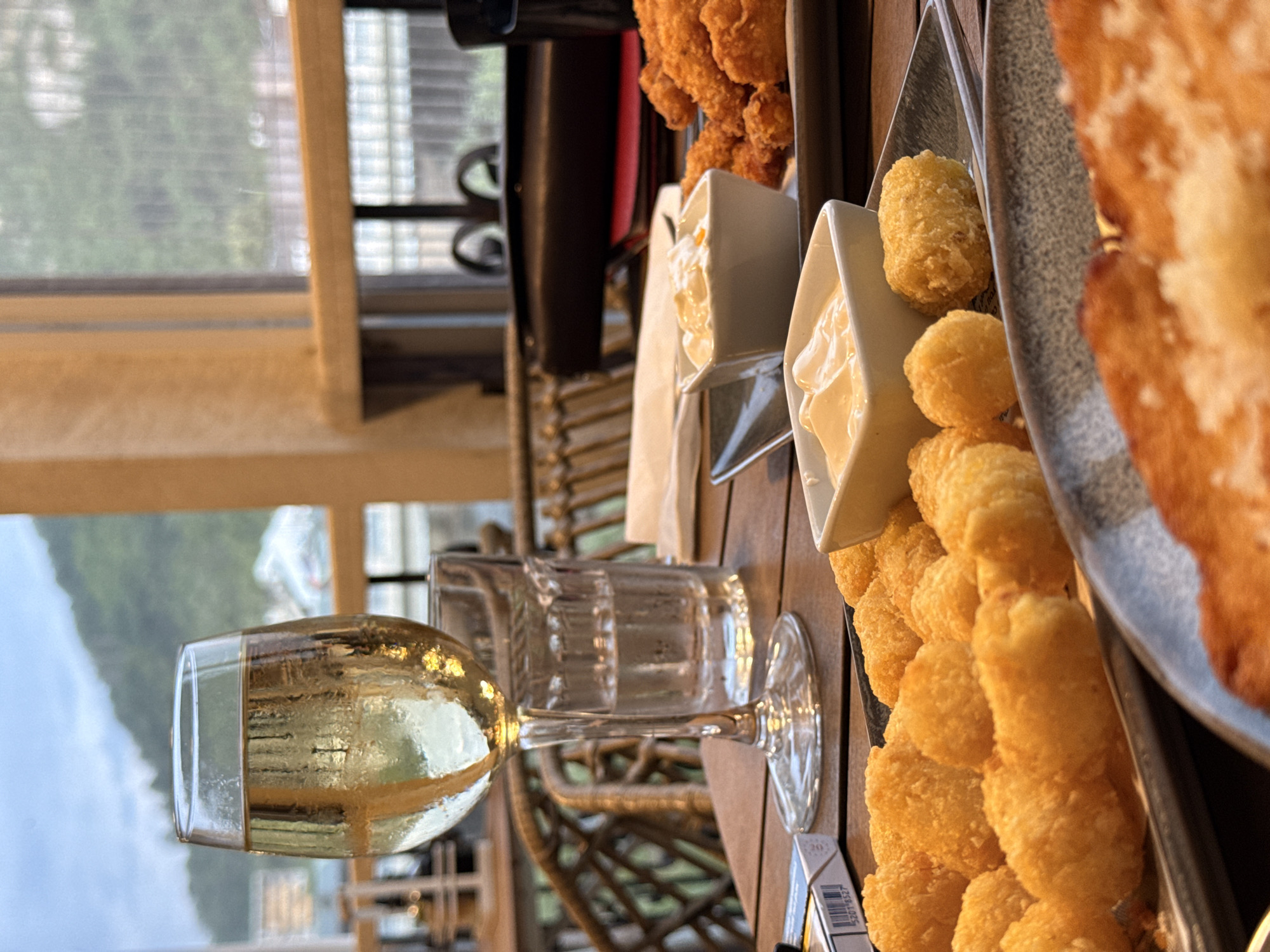 A glass of white wine sits on a wooden table in front of a window overlooking a hillside. In the foreground, a plate of golden-brown fried potato bites and two small bowls of dipping sauce are visible. A pack of cigarettes and a glass of water are also on the table. The scene appears to be a casual dining setting, possibly a restaurant or cafe, with the view suggesting a location in or near Lovech, Bulgaria, given the hilly landscape.