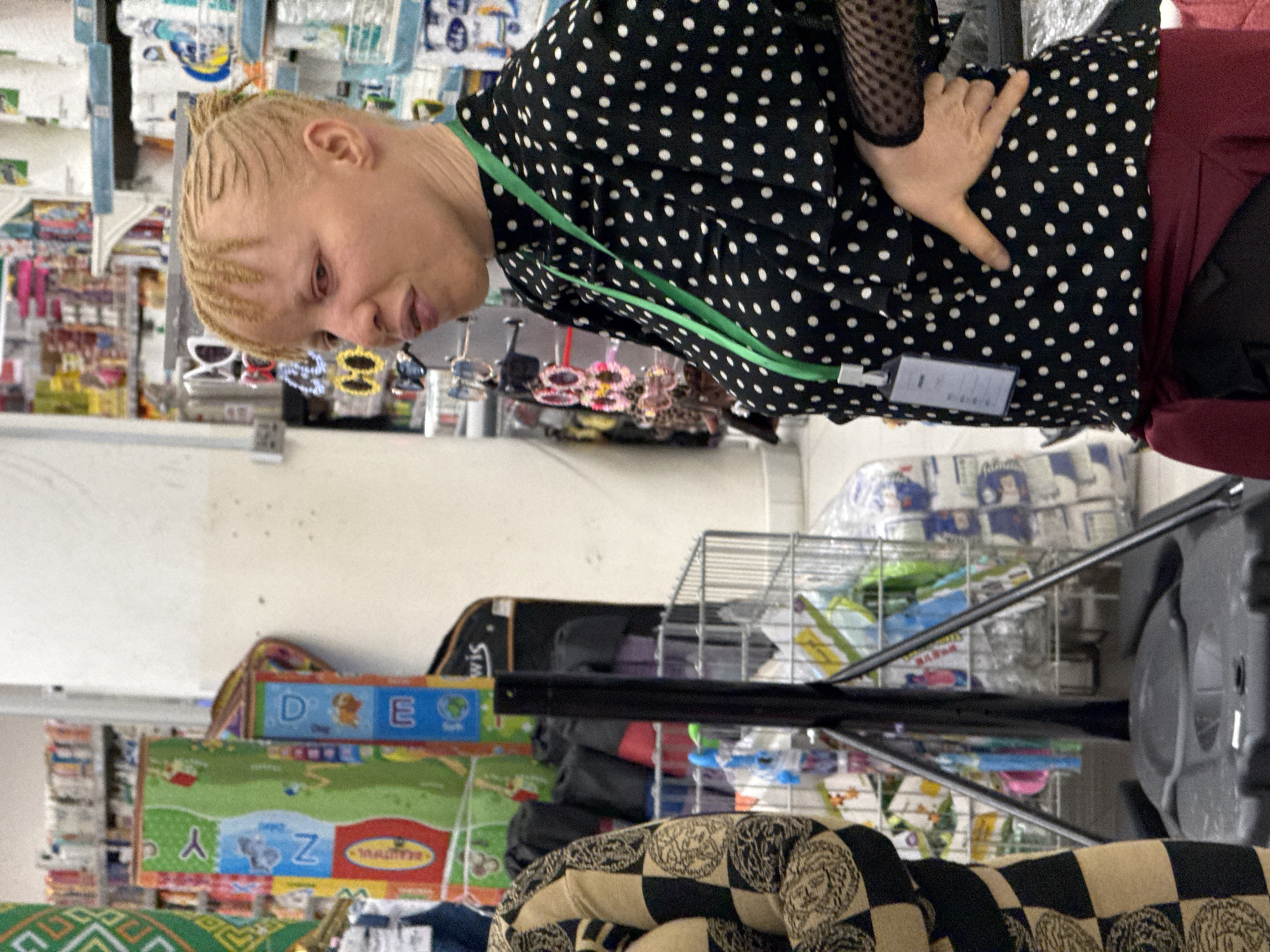 The image shows a person with light blonde braided hair, wearing a black and white polka dot shirt with a green lanyard, standing in a shop with shelves stocked with various goods. The person is looking slightly downward. In the background, there are displays of sunglasses and other items on a shelf. Further back, shelves are filled with packages of what appears to be toilet paper. Below the shelves are children's ABC mats and other items. The location appears to be a retail store, possibly in Abuja, Nigeria.