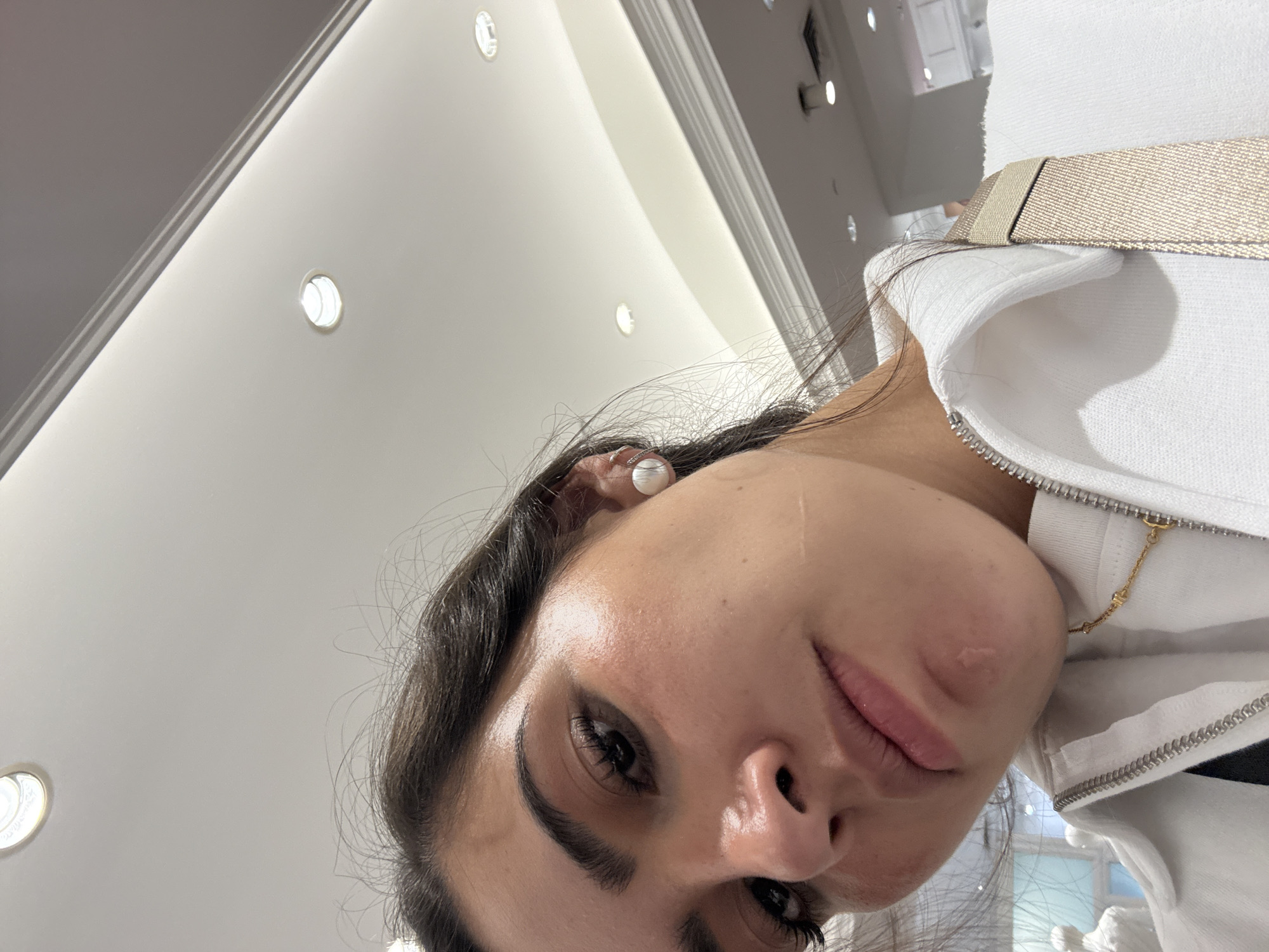 The image is a close-up, slightly tilted selfie of a woman. She has dark hair, dark eyes, and fair skin. She is wearing a white earring and a white jacket with a zipper. There is a beige strap visible near her shoulder. Her facial expression is neutral. The background is out of focus but appears to be an interior space with a white ceiling and recessed lighting.
Without additional information or context, it is impossible to definitively determine the location in San Antonio, United States of America. The image is too close up and lacks identifiable details to discern the specific location.