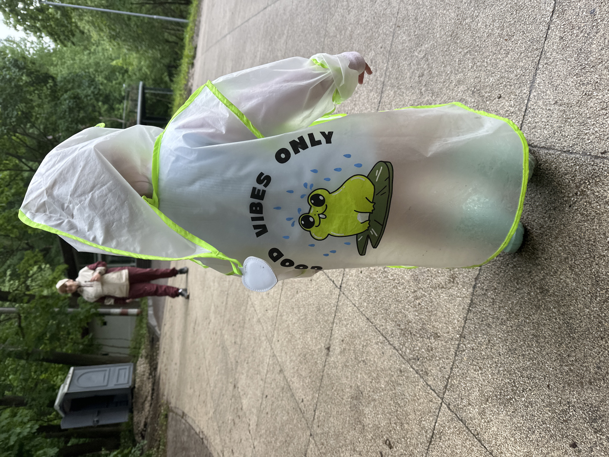 A child wearing a white and clear rain poncho stands on a paved surface. The poncho has neon green trim and a cartoon frog design with the words "Good Vibes Only." A person is walking in the background, and a portable toilet is visible in the distance. The location is a public area, possibly a park or recreational space. The city, Nizhny Novgorod, Russia, is based on the image metadata.