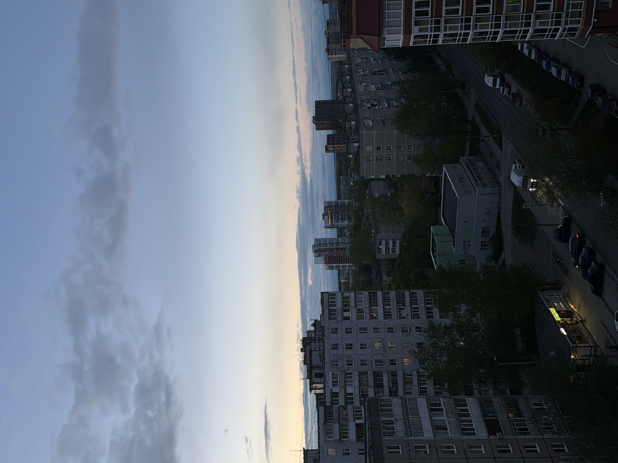 The image captures a cityscape in Perm, Russia, likely taken from a high vantage point such as a building rooftop or upper floor. The view is dominated by several multi-story residential buildings of Soviet-era construction, identifiable by their uniform, rectangular architecture and rows of windows. The buildings are constructed of concrete. A street runs between the buildings. Cars are parked on the side of the street. In the distance, glimpses of other buildings and the skyline are visible. The sky is overcast with clouds.