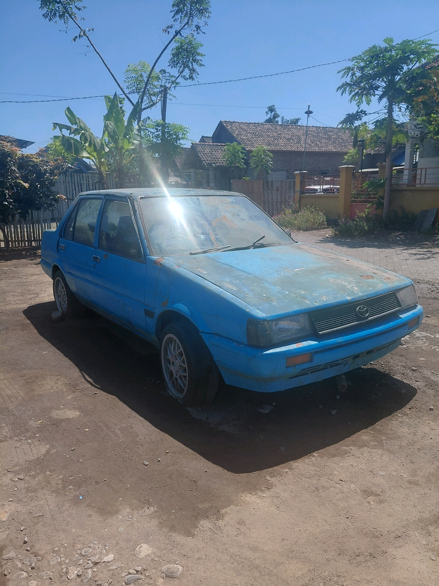 A faded blue, four-door sedan, possibly a Toyota Corolla, is parked on a dirt road under a clear blue sky. The car appears aged and weathered, with visible rust spots and peeling paint on its hood and fenders. The sun is shining brightly, casting a strong shadow to the right of the car. In the background, there are residential buildings with tiled roofs, trees, and a wooden fence. The environment suggests a rural or semi-rural setting, consistent with Paiton, Indonesia. There are no people visible in the image.