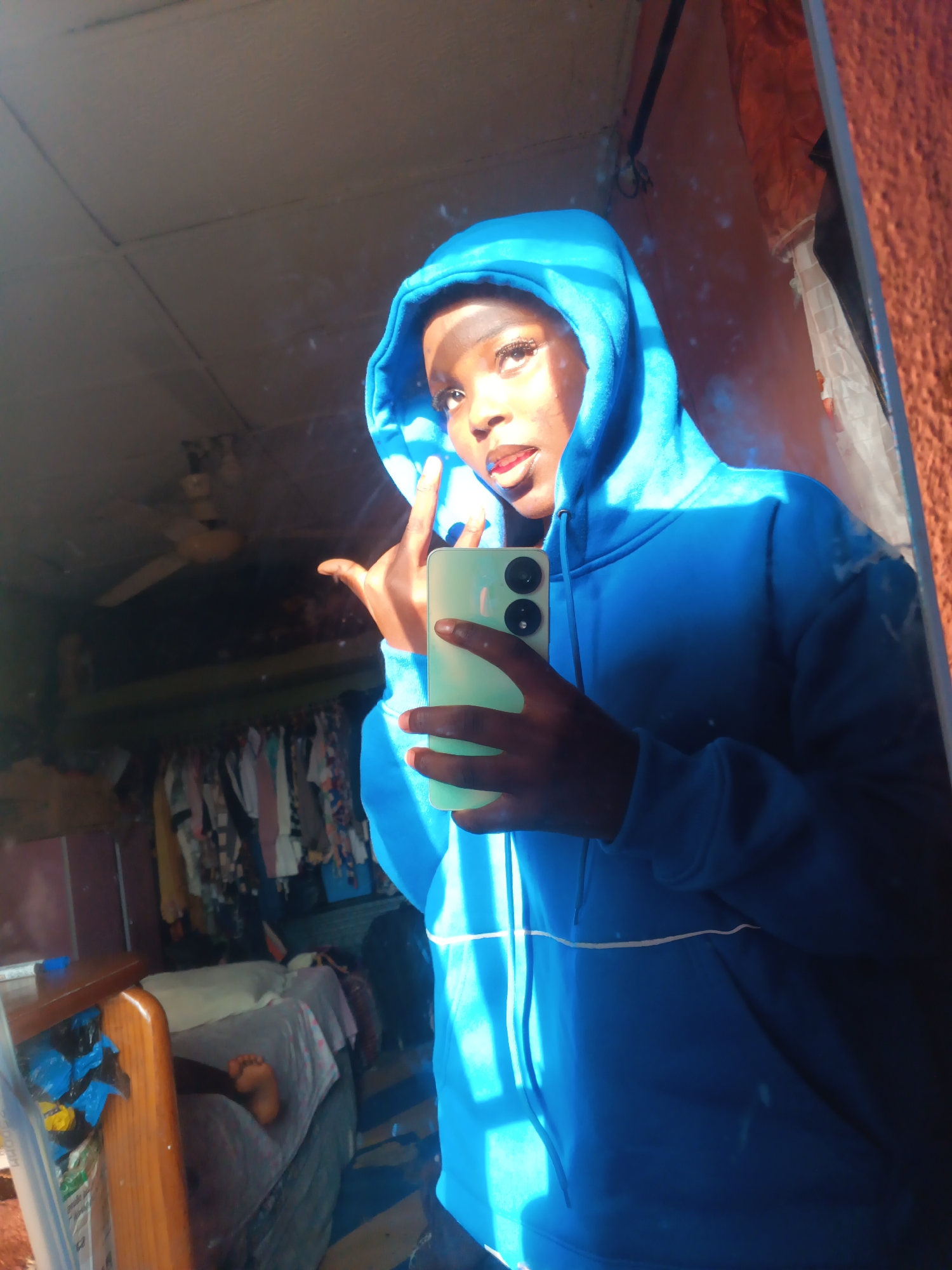 This image depicts a female individual taking a mirror selfie inside a room located in Onitsha, Nigeria. The individual, who has dark skin, is wearing a bright blue hooded sweatshirt with the hood up, partially obscuring the top of their head. They have prominent eyelashes and blue lipstick. Their right hand (viewer's left) holds a light green smartphone with two distinct black circular camera lenses on the back. Their left hand (viewer's right) is raised, making a peace sign (V-sign) gesture. Strong, natural light illuminates the subject's face, front of the hoodie, and parts of the background, creating noticeable glare and reflections on the mirror surface, which also shows some smudges.
The reflection in the mirror reveals a domestic interior. A light-colored ceiling with visible panels is seen at the top. A brown ceiling fan with light fixtures is mounted centrally. To the left, a wooden bed frame with a mattress, a white pillow, and a patterned sheet is visible. A bare foot, likely belonging to another individual, rests on the bed. Below the bed, a blue plastic bag and other items are partially visible. A wooden table or bedside unit is next to the bed. Further back in the room, a clothes rack heavily laden with various garments is visible. The wall to the right of the subject (left in reflection) has a reddish-orange, textured finish. The floor in the lower right corner shows a dark blue surface with light-colored stripes.