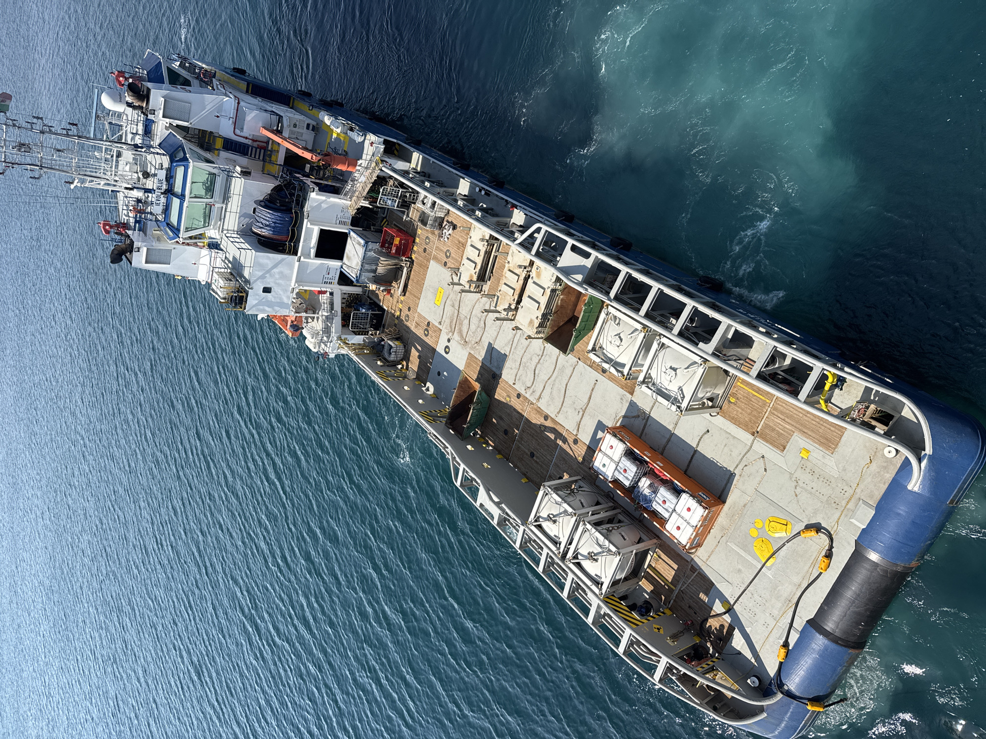 An aerial shot captures a ship with a blue hull at sea, its deck laden with containers and equipment. The ship is positioned at an angle, with its bow pointing towards the upper left of the frame. The water surrounding the vessel is a deep blue, with lighter turquoise patches indicating shallower areas or water disturbance. The sea surface is slightly rippled, suggesting a calm to moderate weather condition.
The ship's deck is mostly a light gray, with wooden sections and yellow markings. Several white, cage-like structures are visible, along with an orange shipping container filled with white cylindrical objects, possibly drums or tanks. A large black and blue cylindrical object is attached to the stern of the ship, extending into the water.
The deck also features various pieces of equipment, including what appear to be cranes, winches, and safety railings. The superstructure of the ship, located towards the front, is white with blue accents and features large windows. Text visible on the side of the superstructure reads "PION BLUE". Another piece of text is visible on the railing, which is difficult to decipher but appears to be a warning or identification.
The scene is illuminated by bright sunlight, casting distinct shadows on the deck and water, indicating it is likely daytime. The overall impression is one of industrial activity at sea, likely involved in cargo transport or offshore operations. No people are visible in the frame, focusing the attention on the vessel and its cargo.