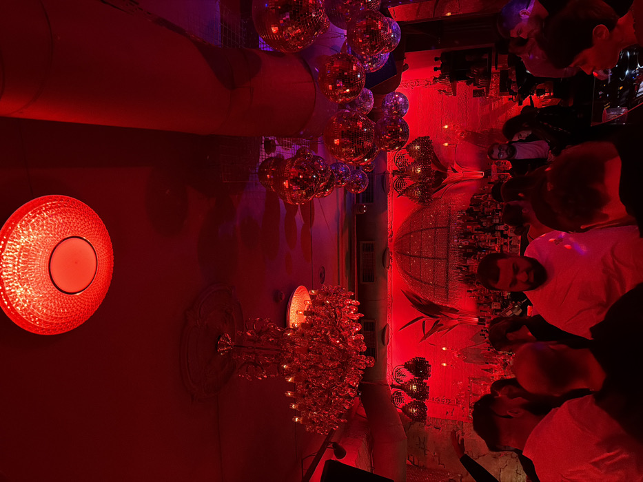 A bustling bar scene is awash in vibrant red lighting. Several disco balls hang from the ceiling, reflecting the light around the room. A large, ornate chandelier adds to the ambiance. Patrons cluster around the bar, some facing the camera, their faces partially obscured. A circular red light fixture on the wall provides further illumination. The overall impression is one of a lively and crowded nighttime establishment.