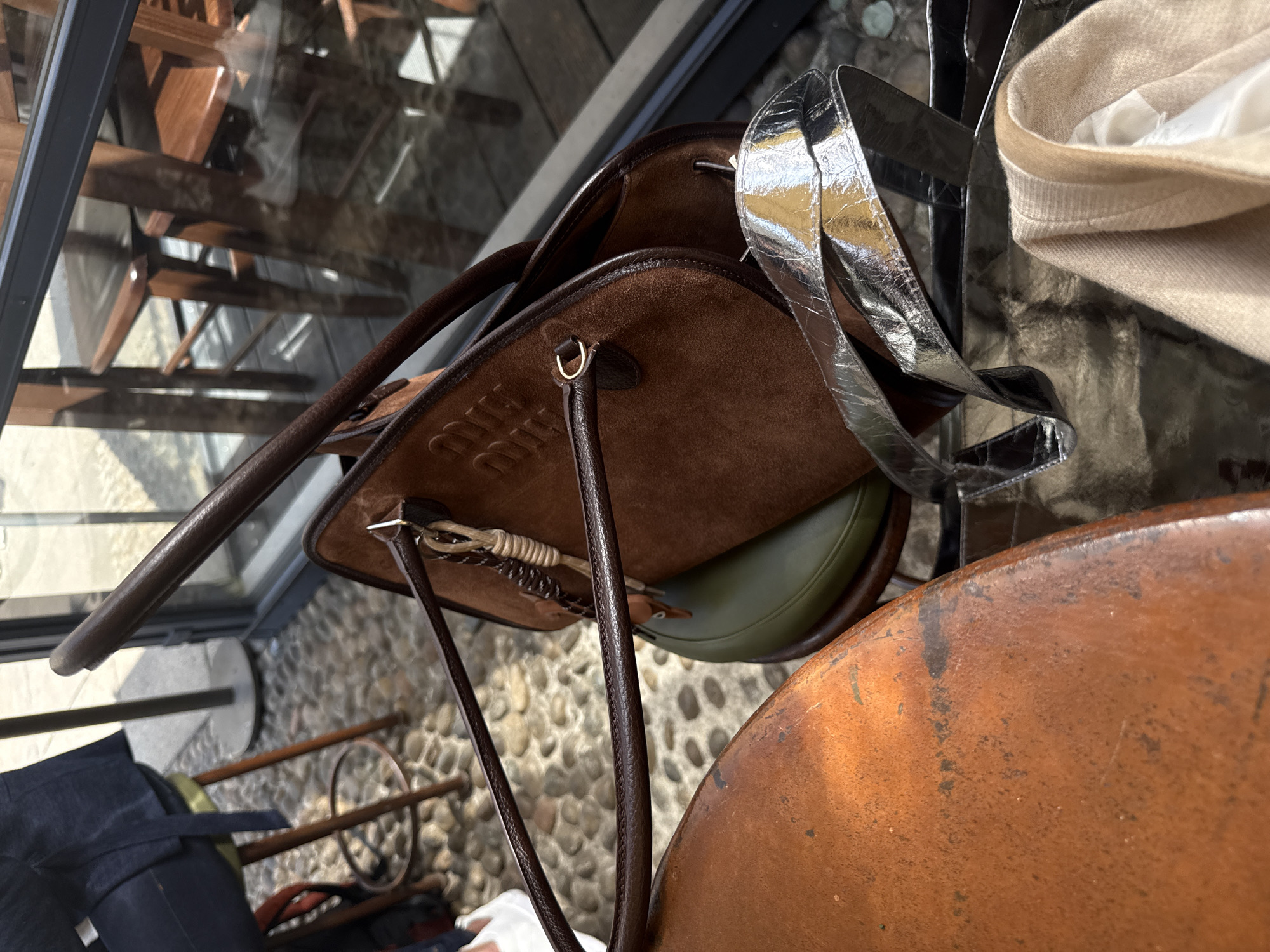 The image, rotated counter-clockwise, presents a close-up, downward view of several fashion accessories arranged on a reflective glass surface.
In the foreground and central, a prominent brown, suede-like bag features dark brown leather trim and handles. The brand name "Miu Miu" is subtly embossed on its front, and a small, gold-colored D-ring is visible. Partially seen behind this, an olive green-colored bag adds a hint of contrasting color. To the right, a crinkled, silver metallic material, possibly a bag or a strap, reflects light. Adjacent to the silver item, a light beige or cream-colored knitted garment, likely a sweater or scarf, is neatly folded. The bottom right corner shows a segment of a worn, reddish-brown, textured surface, resembling an aged wooden table or stool.
The glass surface reflects abstract wooden architectural elements, possibly shelving or stairs, suggesting an indoor setting with modern interior design. Through the glass, the floor below is visible, featuring a distinct cobbled or pebbled pattern. On this floor, glimpses of other objects are discernible, including dark blue fabric items and slender wooden or metal rods, hinting at additional displays or furnishings.
The scene suggests an arrangement of fashion items, likely within a boutique, showroom, or high-end retail environment in Milan, presented as a display. No human activity is depicted. The lighting appears soft and indirect, characteristic of an indoor setting. The visible text is "Miu Miu" on the brown bag.