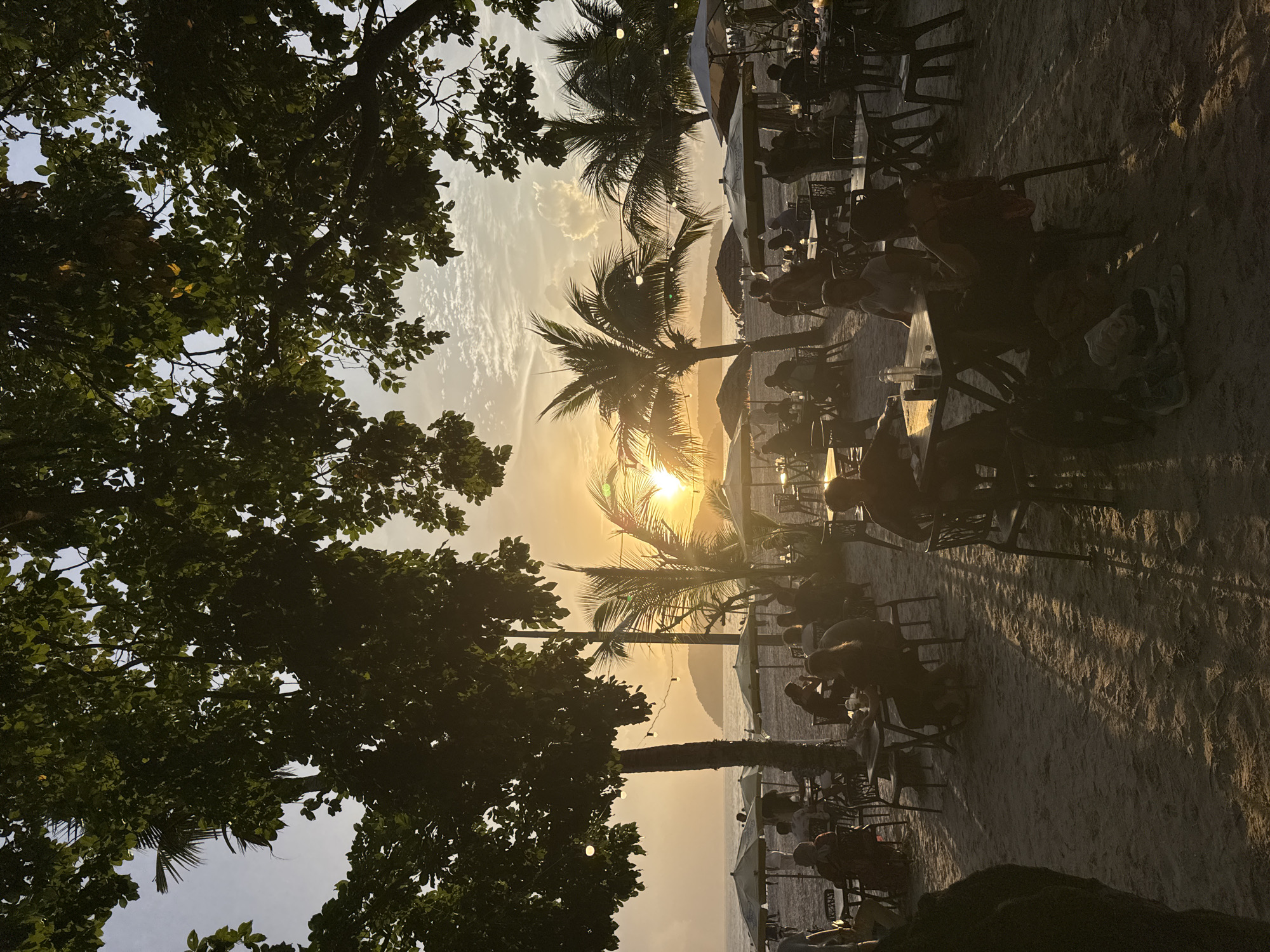 This image depicts a beachside dining scene at Cenang Beach, Malaysia, during sunset. The foreground features several tables and chairs set up on the sandy beach, occupied by patrons. These individuals are silhouetted against the warm glow of the setting sun. In the background, palm trees frame the horizon, and the ocean is visible with a distant island or landmass. Umbrellas are set up over some of the tables, providing shade. String lights are visible hanging from trees, suggesting a leisurely evening atmosphere. The sun is low in the sky, casting long shadows across the sand and creating a golden hour ambiance. The sky is partly cloudy, with the sun radiating a bright light.