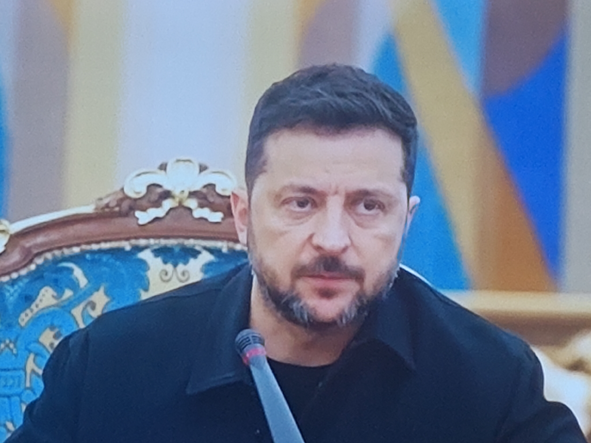 The image shows a head and shoulders portrait of a man wearing a black jacket over a black t-shirt. He has short, dark hair, a beard, and a mustache. His eyes are open and looking forward. He is seated in front of a microphone. The background is blurred and appears to be an indoor setting with abstract geometric shapes in shades of blue and yellow. The man's left shoulder and the back of his neck are visible, as is part of an ornate chair with blue upholstery and carved wooden details. The image quality is low, with visible pixelation and noise. There is no information in the image to suggest a location in Molenbeek-Saint-Jean - Sint-Jans-Molenbeek, Belgium.
