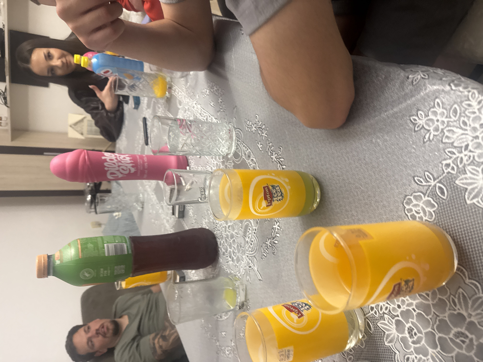 The image shows a gathering of people around a table covered with a gray tablecloth with a white floral pattern. The table holds several glasses and bottles, including a green and brown bottle, two clear glasses, a pink bottle, and three yellow Lipton Ice Tea glasses, filled with a yellow liquid. Three individuals are visible: a woman pouring a blue bottle of liquid into a glass, a man in a green shirt is visible in the bottom left corner, and the arm and shoulder of another person are in the upper right corner.
The setting appears to be indoors. There is no verifiable evidence to confirm this location is Graf Ignatievo, Bulgaria.