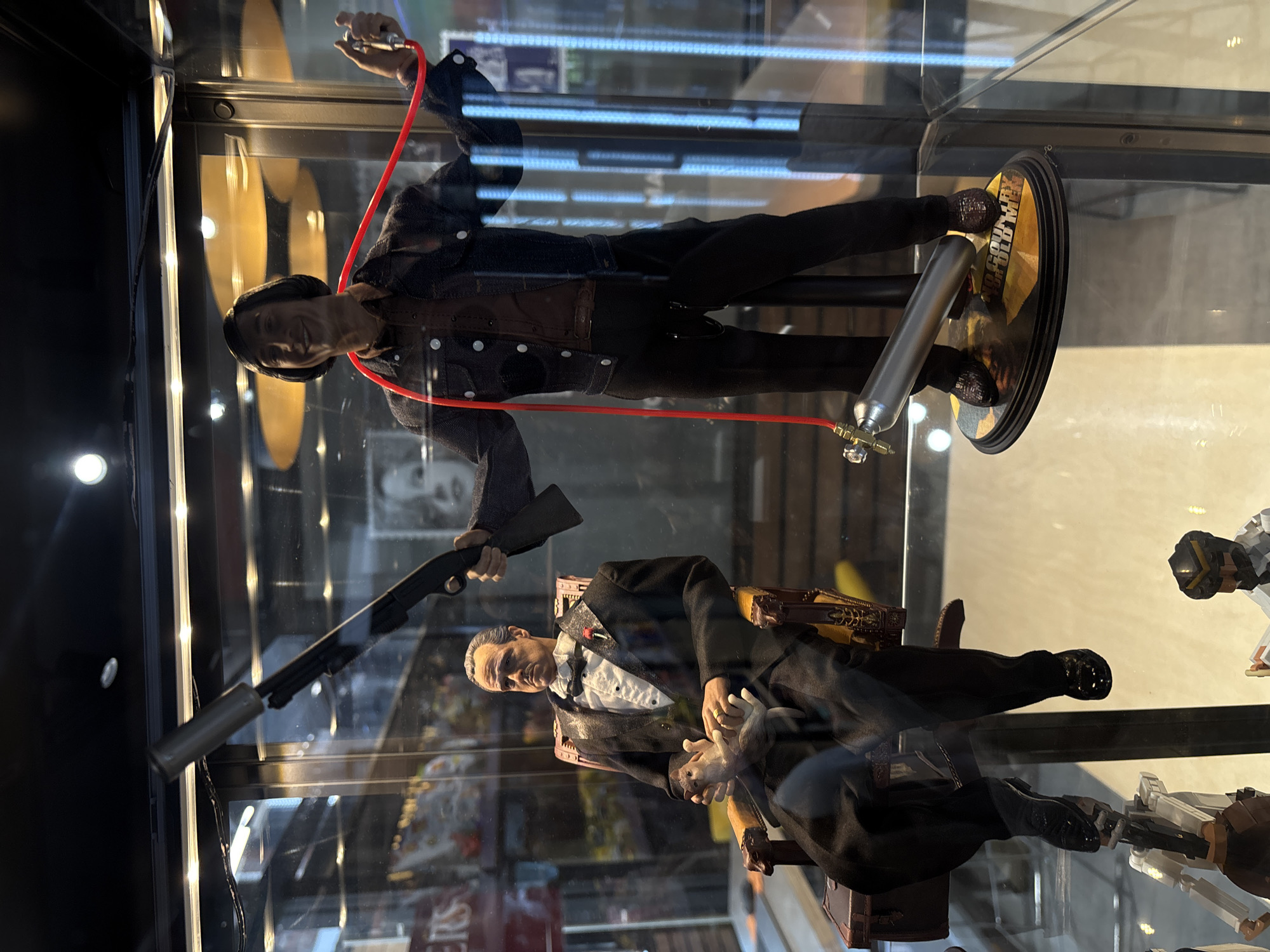 The image depicts a display case containing action figures. At the top, a figure resembling Anton Chigurh from "No Country for Old Men" stands with a captive bolt pistol and an oxygen tank. The display base indicates the film title. Below, a figure resembling Vito Corleone from "The Godfather" sits in a chair, holding a white cat. Another figure holds a shotgun aimed towards Corleone. Additional figures are visible in the lower right of the display. The location appears to be a retail setting, possibly a store specializing in collectibles.