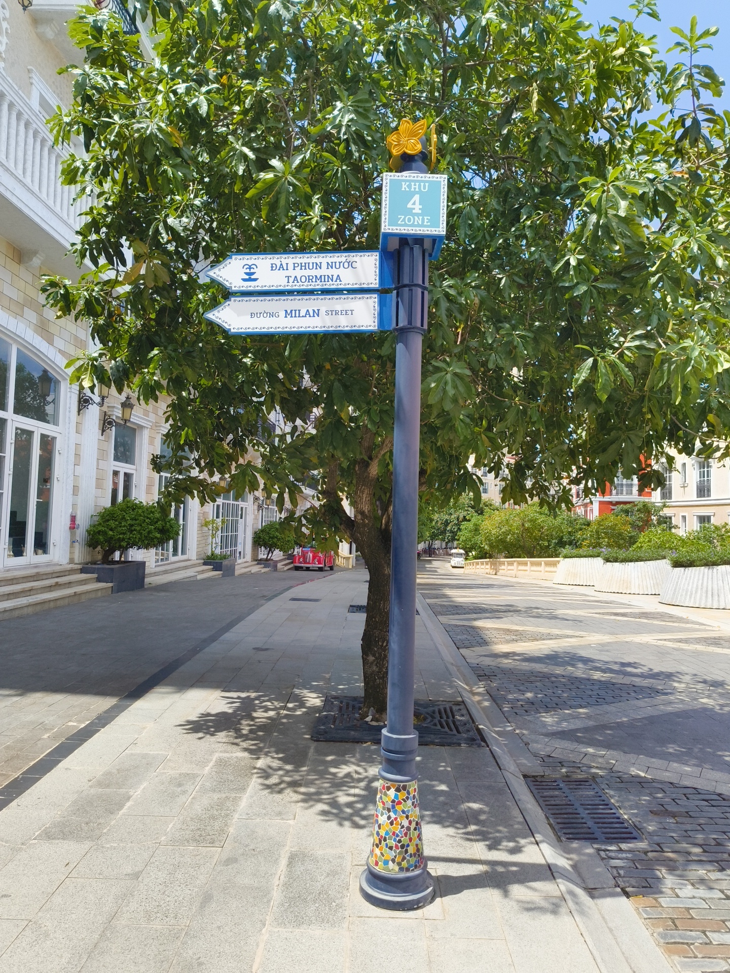 A signpost stands on a paved walkway in front of a building with large windows. The signpost has multiple directional signs and a square sign with "KHU 4 ZONE" written on it. One sign points left and reads "ĐẠI PHUN NƯỚC TAORMINA". Another sign points left and reads "ĐƯỜNG MILAN STREET". The signpost is decorated with colorful mosaic tiles at its base. A tree with green leaves partially obscures the signpost. The building is multi-story with white balconies and tan-colored walls. The walkway is made of gray pavers. In the background, there are other buildings and foliage. A red vehicle is visible further down the street. The scene is outdoors on a sunny day. The sign indicating "ĐƯỜNG MILAN STREET" suggests the location is in Phú Quốc, Vietnam, as "Đường" is the Vietnamese word for "street".