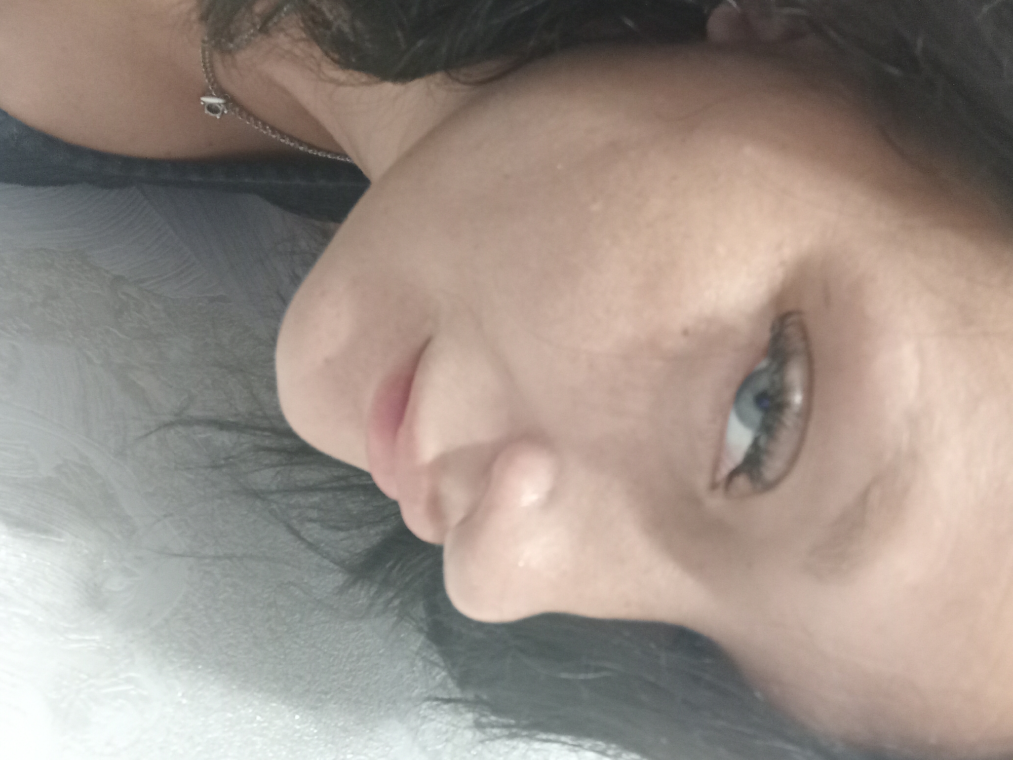 The image shows a close-up, angled selfie of a young woman with dark hair and blue eyes. She is wearing a necklace with a small charm. Her face is partially obscured by her hair, and she is looking slightly downwards and to her left. The background appears to be a textured, light-colored wall. No specific location in Chuhuiv, Ukraine is identifiable from the image.