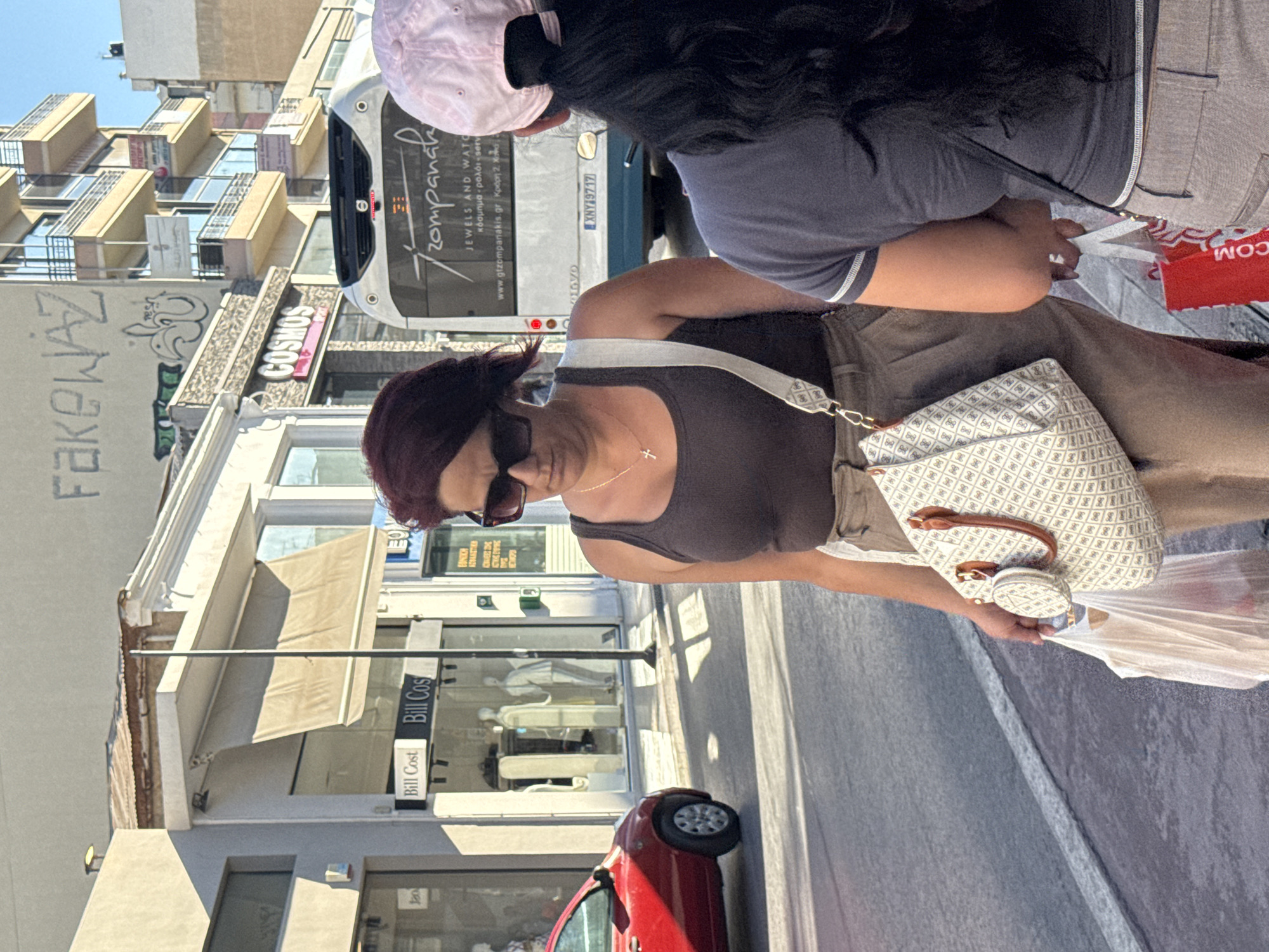 The image depicts a street scene in Chania, Greece, likely on a sunny day. In the foreground, a woman with dark, shoulder-length hair and dark sunglasses is walking. She is wearing a dark tank top and light brown pants. She carries a light-colored patterned handbag with brown leather straps and a small matching circular coin purse. In her left hand, she holds a white plastic shopping bag. To her right, another person with long black hair, wearing a pink baseball cap and a dark t-shirt, is also partially visible. They are holding a red shopping bag. In the midground, a red car is parked on the street. Behind the people, a bus with the text "Tzompanakis" and "JEWELS AND WATCHES" is visible. The building facade behind the bus has the word "COSMOS" in large letters. The buildings in the background appear to be multi-story structures with balconies. Graffiti is present on the upper part of one of the buildings. The street surface is asphalt with white markings.
