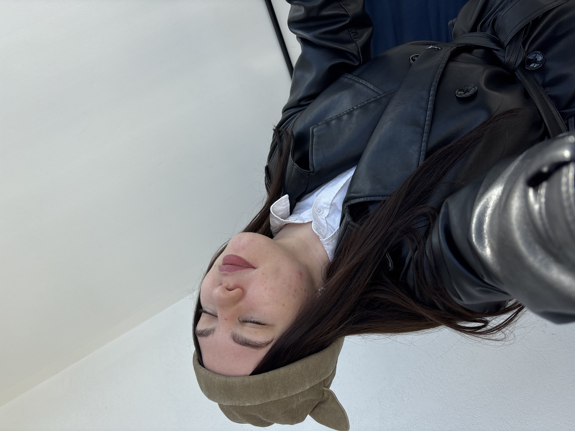 A single young adult female is depicted in a close-up, upward-angled shot, her head tilted back and to the viewer's left, with eyes closed and lips slightly parted. She has long dark hair and is wearing a khaki-colored soft cap, a white collared shirt, and a black faux leather trench coat with a belted waist. Minor skin blemishes are visible on her cheeks and chin. The background is a plain, light-colored surface, likely a wall or ceiling, forming a diagonal line, with an indistinct dark object partially visible on the far right. The lighting is bright and appears even. The specific location within Perm, Russia, is not identifiable from the visible details.