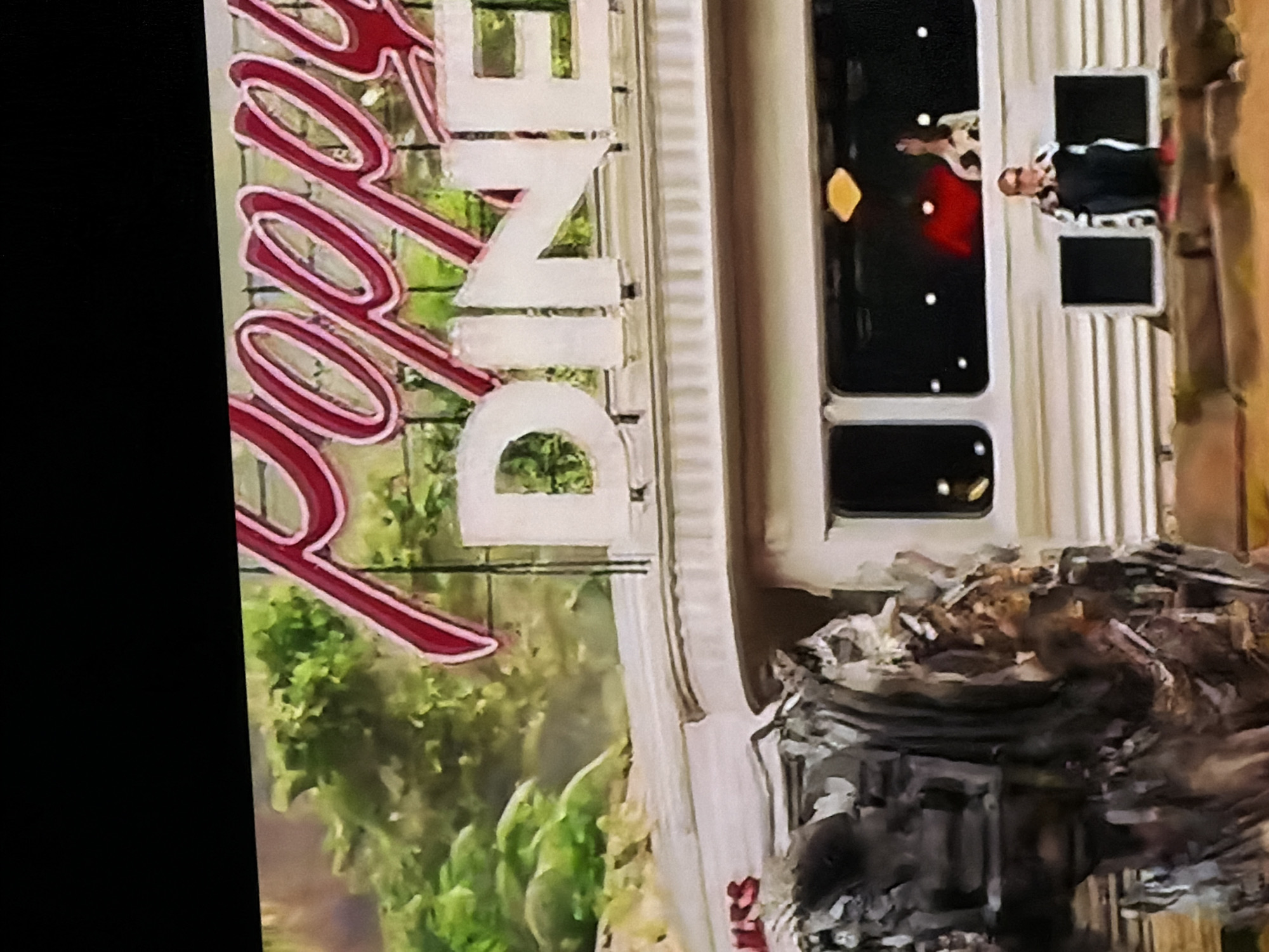 The image, correctly oriented, shows the exterior of a building, identified as "Poppy's Diner" by the prominent sign. The sign features "Poppy's" in red, illuminated cursive script, with "DINE" in large white block letters underneath. The time appears to be dusk or night, indicated by the lit sign and interior lights visible through the windows.
In the upper-right window, a person is visible, looking out and raising their right arm. They are wearing a light-colored, possibly patterned top and dark bottoms. Another, less distinct figure is seated inside near an adjacent window. The windows reflect internal lights.
A notable and critical detail is the significant amount of wreckage and debris accumulated in the foreground, directly below the diner's windows. This suggests damage to the building or its immediate vicinity. Green foliage, likely trees or bushes, can be seen in the background behind the diner sign. The scene, located in Sofiivska Borshchahivka, Ukraine, juxtaposes the functioning diner sign with visible structural damage.