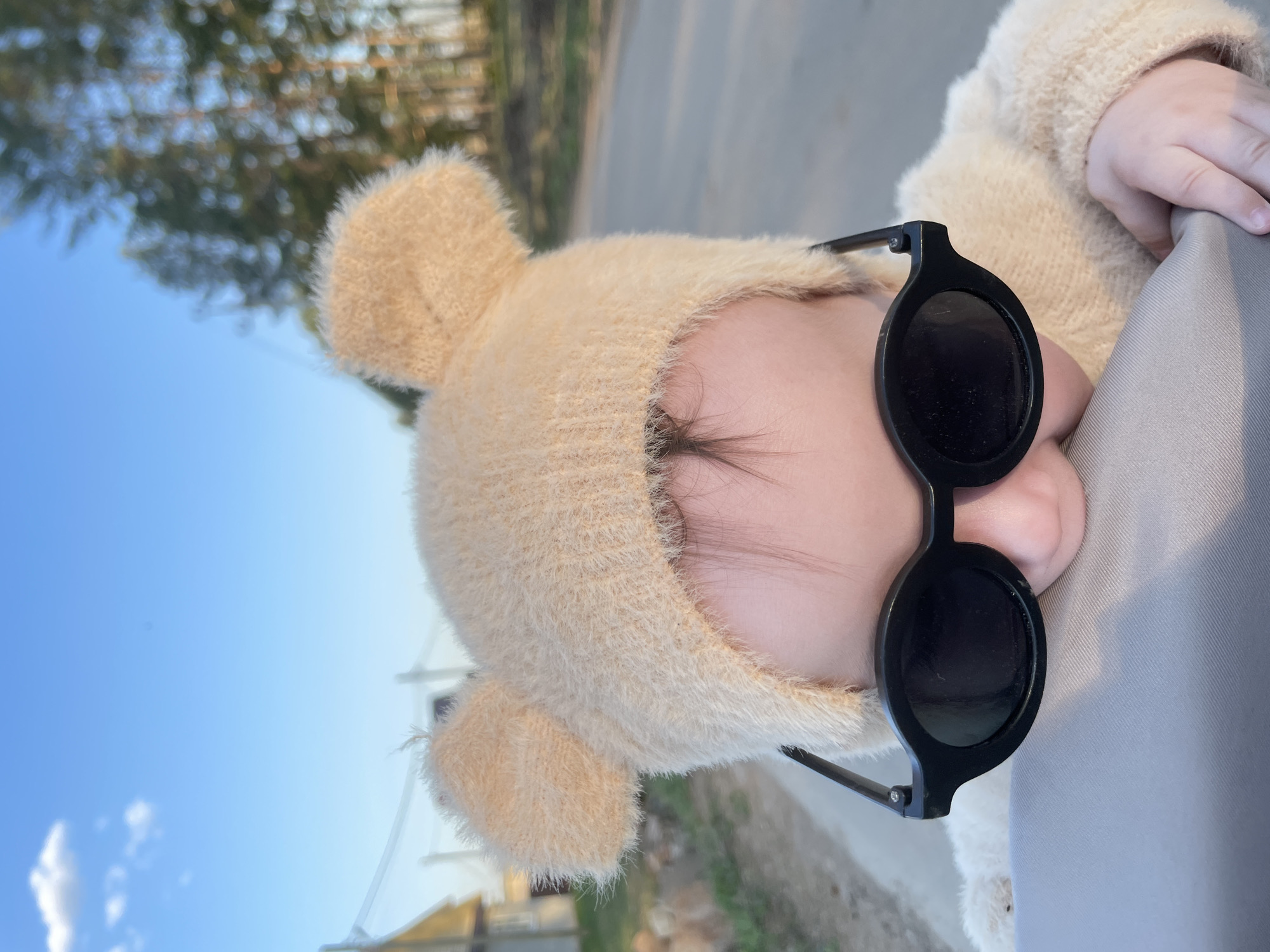 A close-up, canted shot features a child wearing a fuzzy, beige hat with ears and black, round sunglasses. The child's face is largely obscured by the sunglasses, though a glimpse of eyelashes and skin is visible beneath the frame. A small hand rests on a light gray surface in the upper right.
The background is blurred, showing a partly cloudy blue sky and indistinct green foliage with hints of buildings. The specific location is Подшивалово, Russia, as requested.