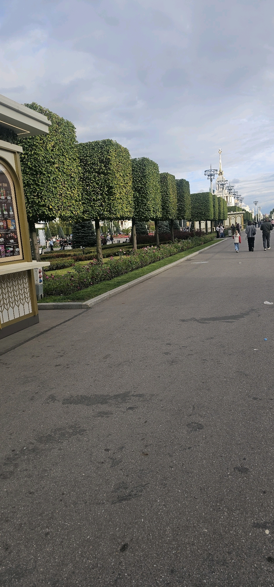 The image shows a paved pathway lined with neatly trimmed trees and flowerbeds. In the background, several people are walking along the pathway. Further in the distance, there are ornate buildings with spires, characteristic of Soviet-era architecture. The sky is overcast with clouds. The scene is consistent with the VDNKh (Exhibition of Achievements of National Economy) park in Moscow, Russia, a large complex known for its pavilions, fountains, and green spaces.