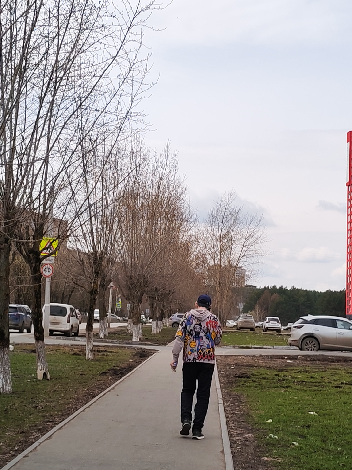 A person in black pants and a colorful patterned jacket walks away from the camera on a concrete sidewalk. The individual is wearing a dark baseball cap. To the left and right of the sidewalk is a strip of grass and bare-branched trees. Several parked cars are visible on the left side of the sidewalk. In the background, there are more trees, a partially visible tall red building, and a sky with scattered clouds. The image appears to be taken in the city of Izhevsk, Russia, based on the presence of characteristic architecture and signage. A "40" speed limit sign with a pedestrian crosswalk sign above it is partially visible on the left side of the image.